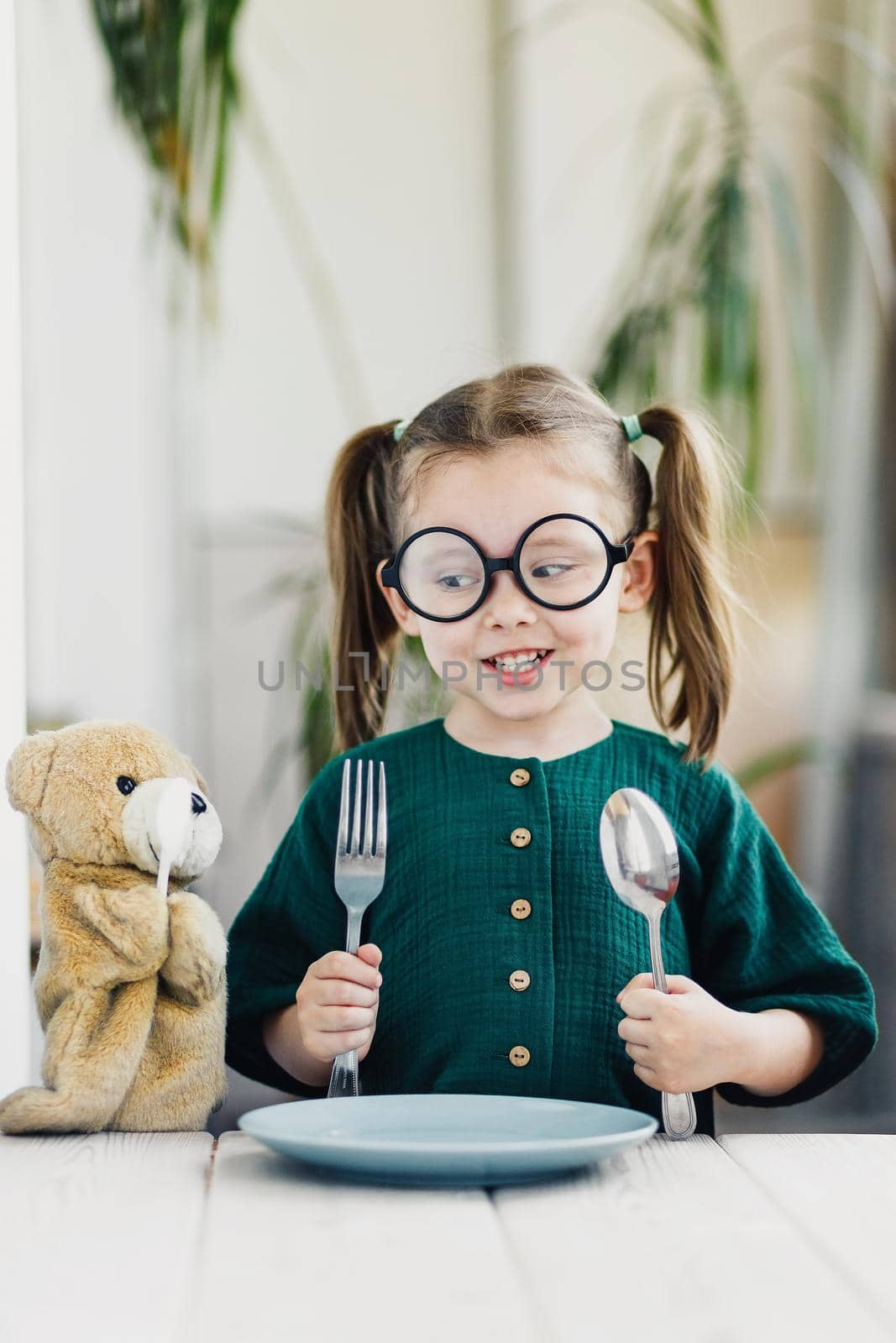 Little beautiful girl in green muslin dress waiting breakfast with her friend bear toy. Little cute girl at white dining table in kitchen. Healthy nutrition for young kids.