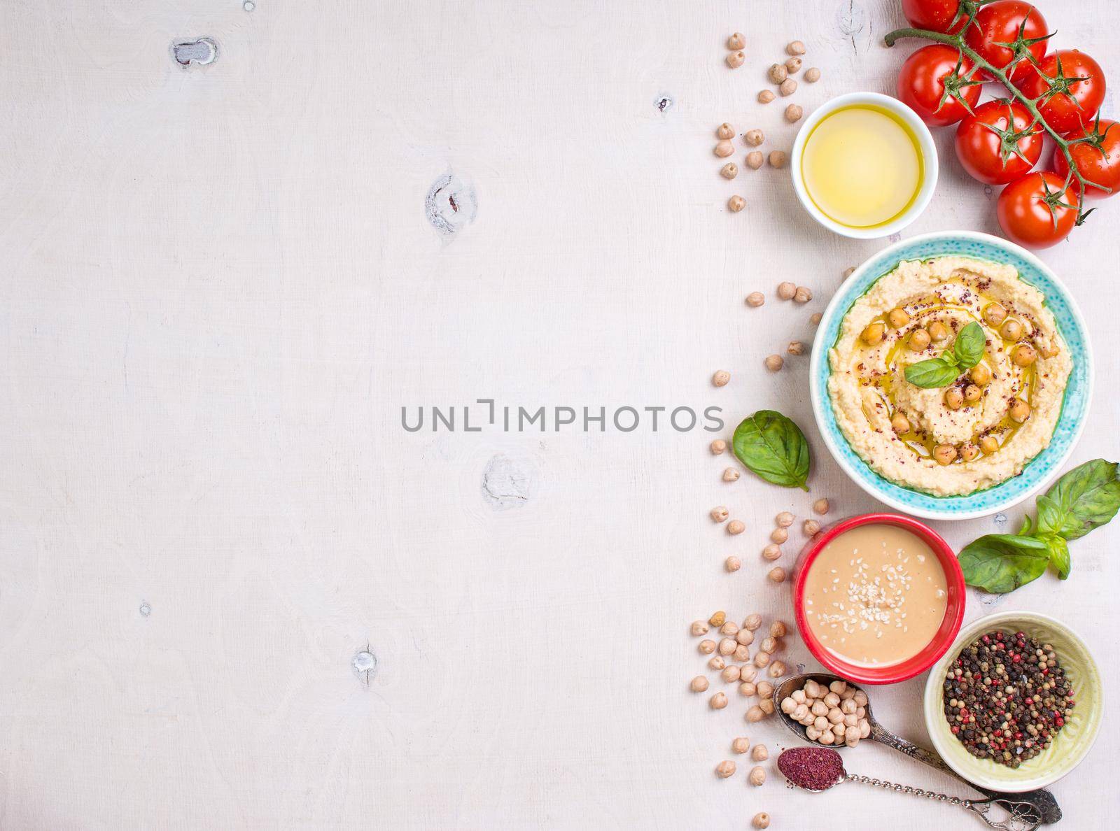 Bowl with hummus, chickpea, tahini, olive oil, sesame seeds, cherry tomatoes and herbs on white rustic wooden background. Space for text. Food frame. Middle eastern cuisine. Top view. Hummus background