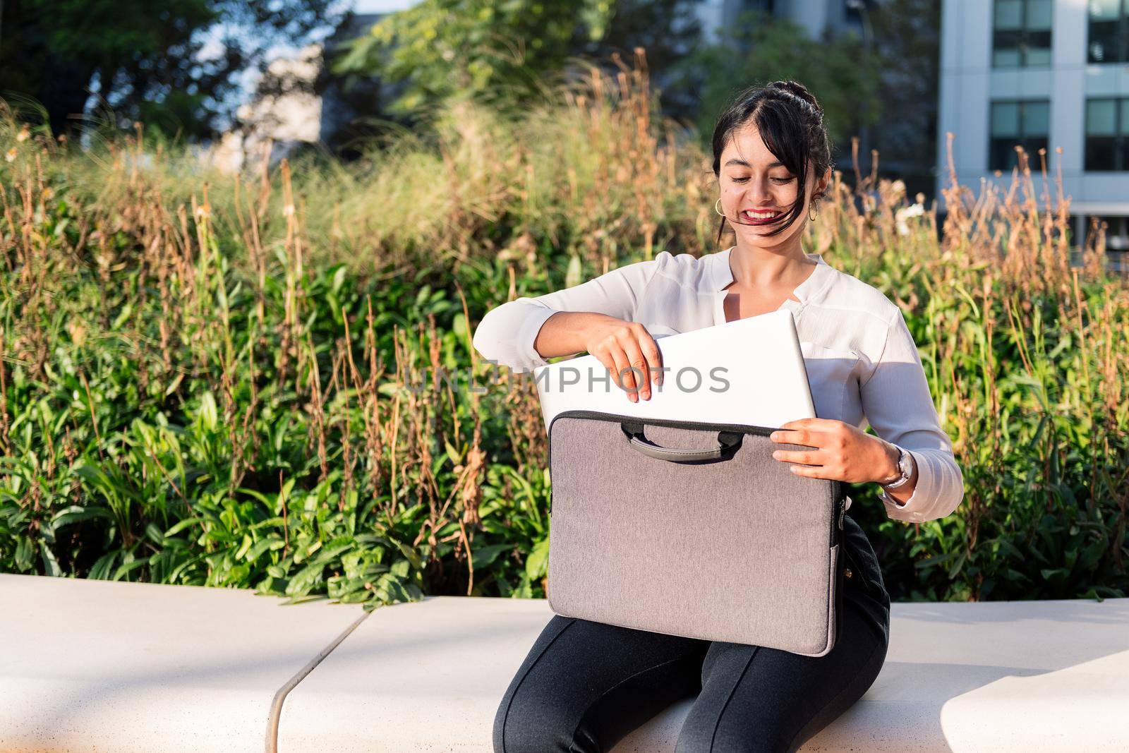 smiling businesswoman taking her laptop computer out of her briefcase sitting in a park, concept of digital entrepreneur and urban lifestyle, copy space for text
