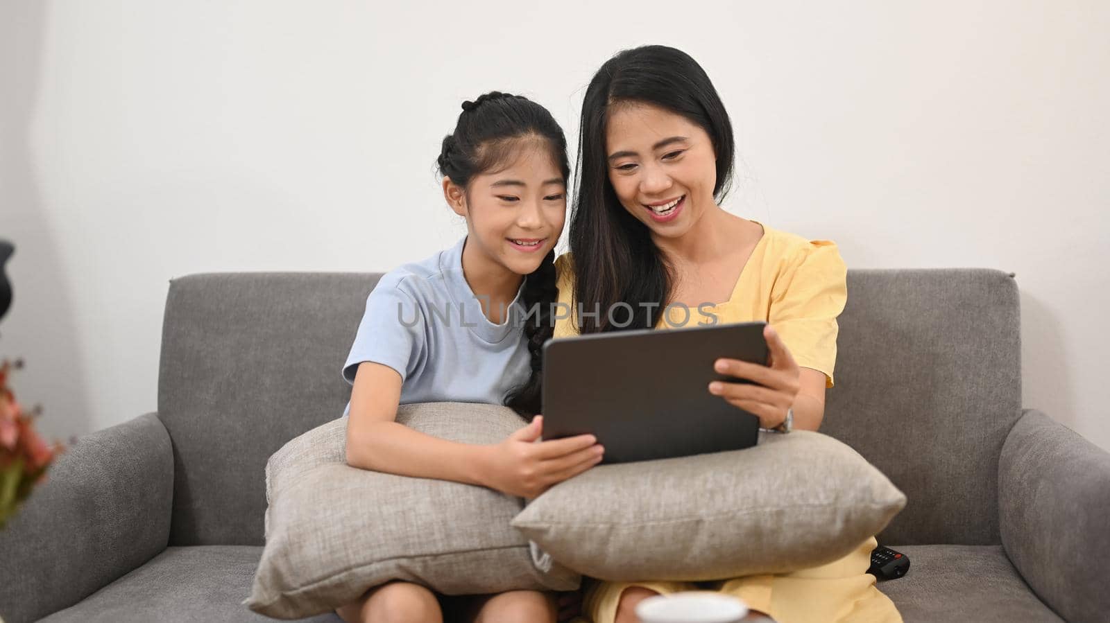 Asian mother and daughter using digital tablet while sitting on sofa, enjoying spending free time together at home by prathanchorruangsak