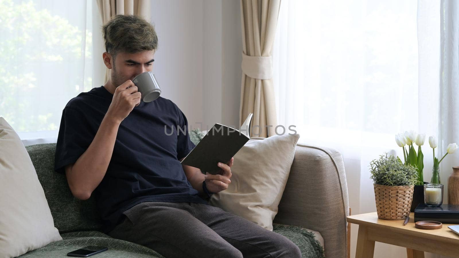 Relaxed millennial man drinking hot coffee and reading book on comfortable sofa by prathanchorruangsak