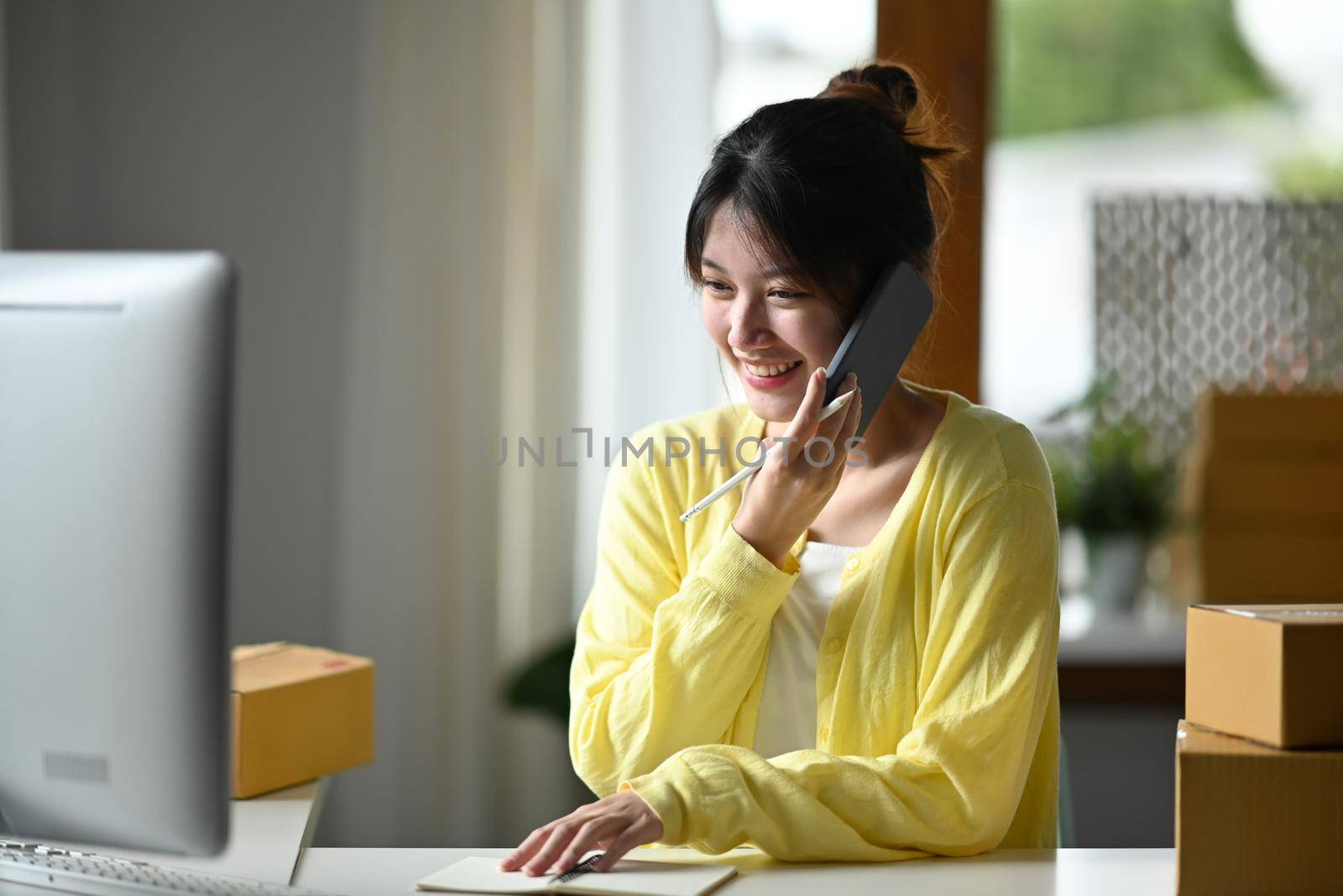 Smiling female small business owner using computer and talking on mobile phone for confirming orders from customer. Online selling, e-commerce concept.