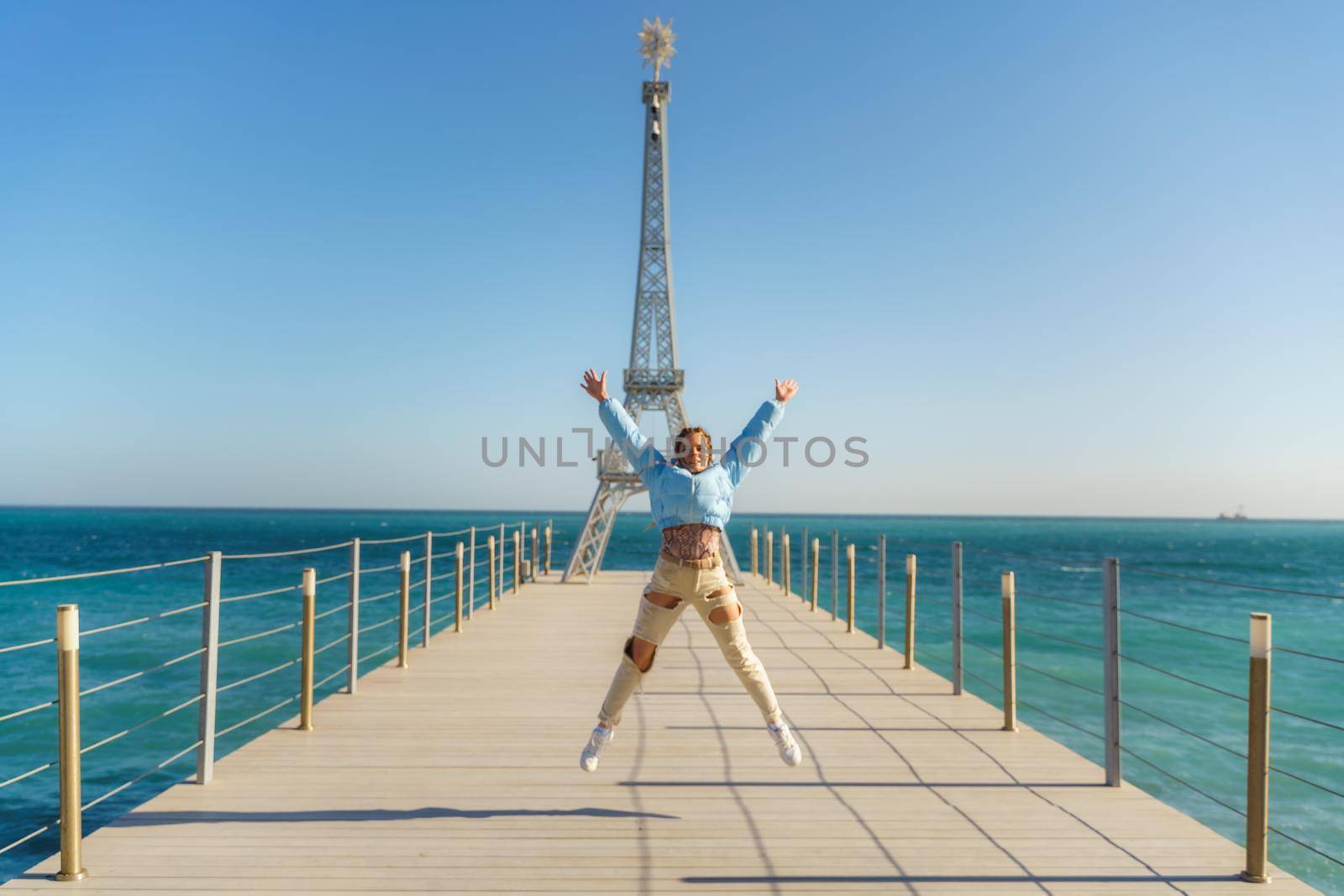 Large model of the Eiffel Tower on the beach. A woman walks along the pier towards the tower, wearing a blue jacket and white jeans. by Matiunina