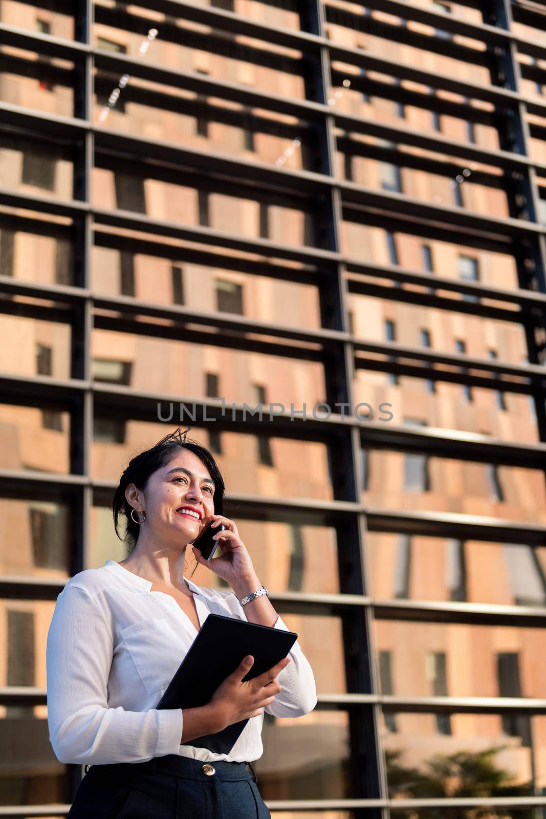smiling businesswoman talking on the phone and using her tablet in front of an office building, concept of successful entrepreneur and urban lifestyle, copy space for text