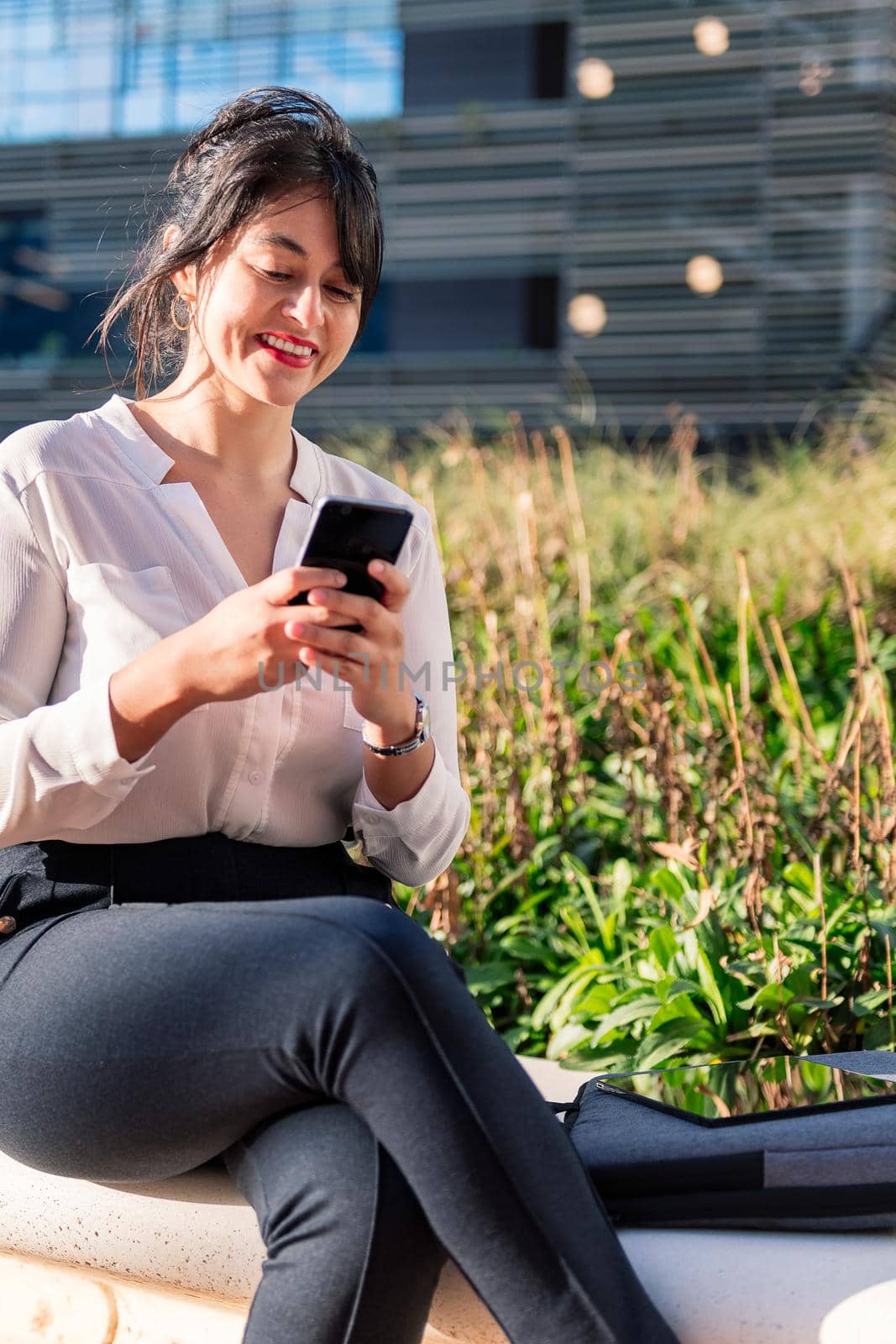 smiling businesswoman typing on her phone sitting in a park in the financial district, concept of digital entrepreneur and urban lifestyle, copy space for text