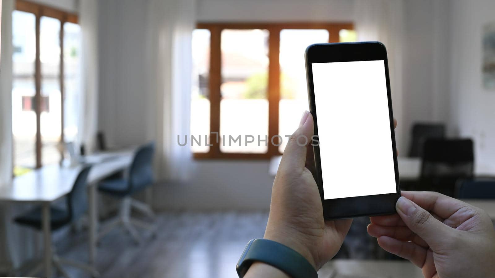 Close up view of man hands holding mock up smart phone with blurred office background in morning light.