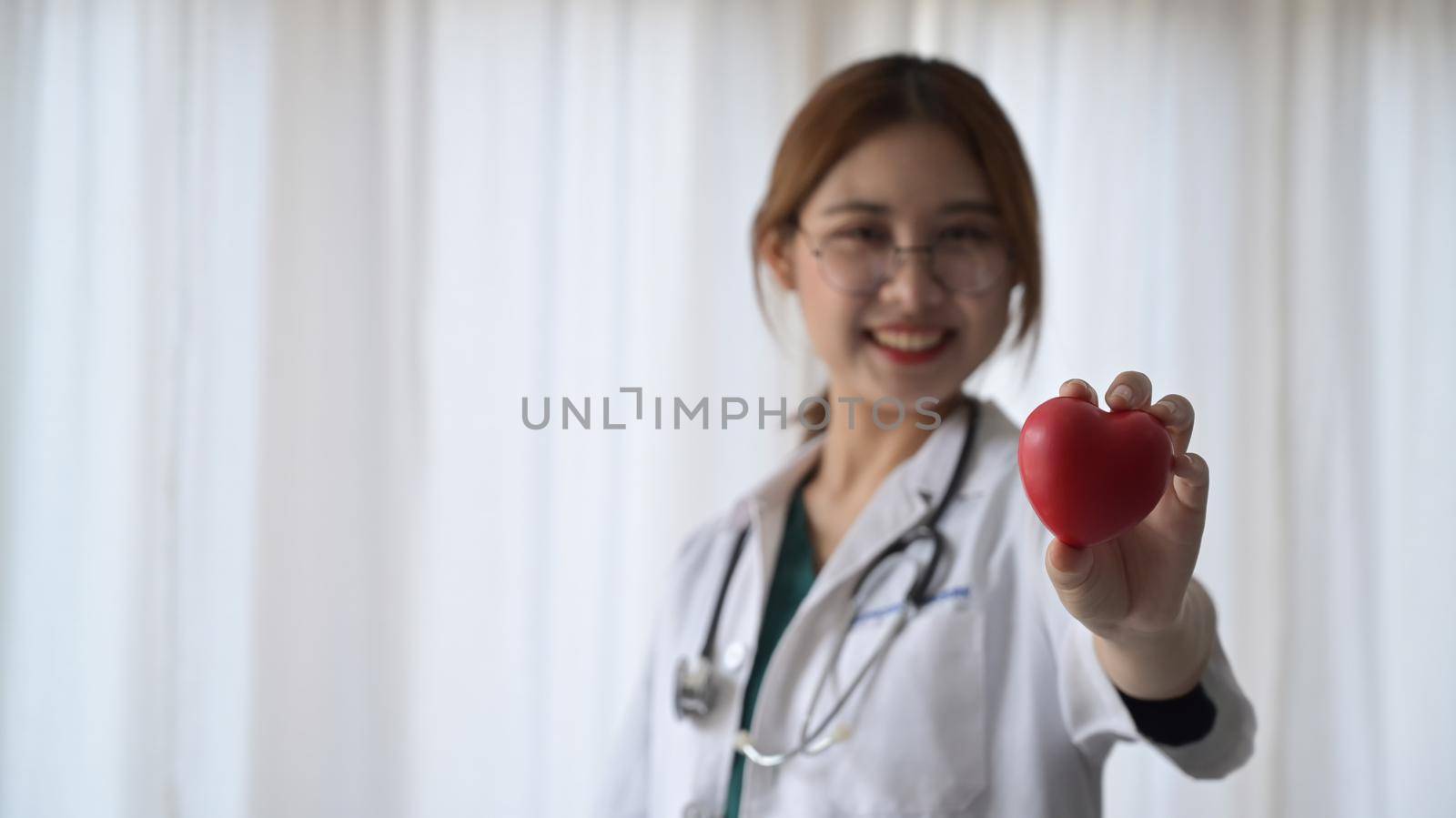 Smiling female doctor in white medical uniform holding red heart shape in hand. Cardiology, healthcare and medical concept.