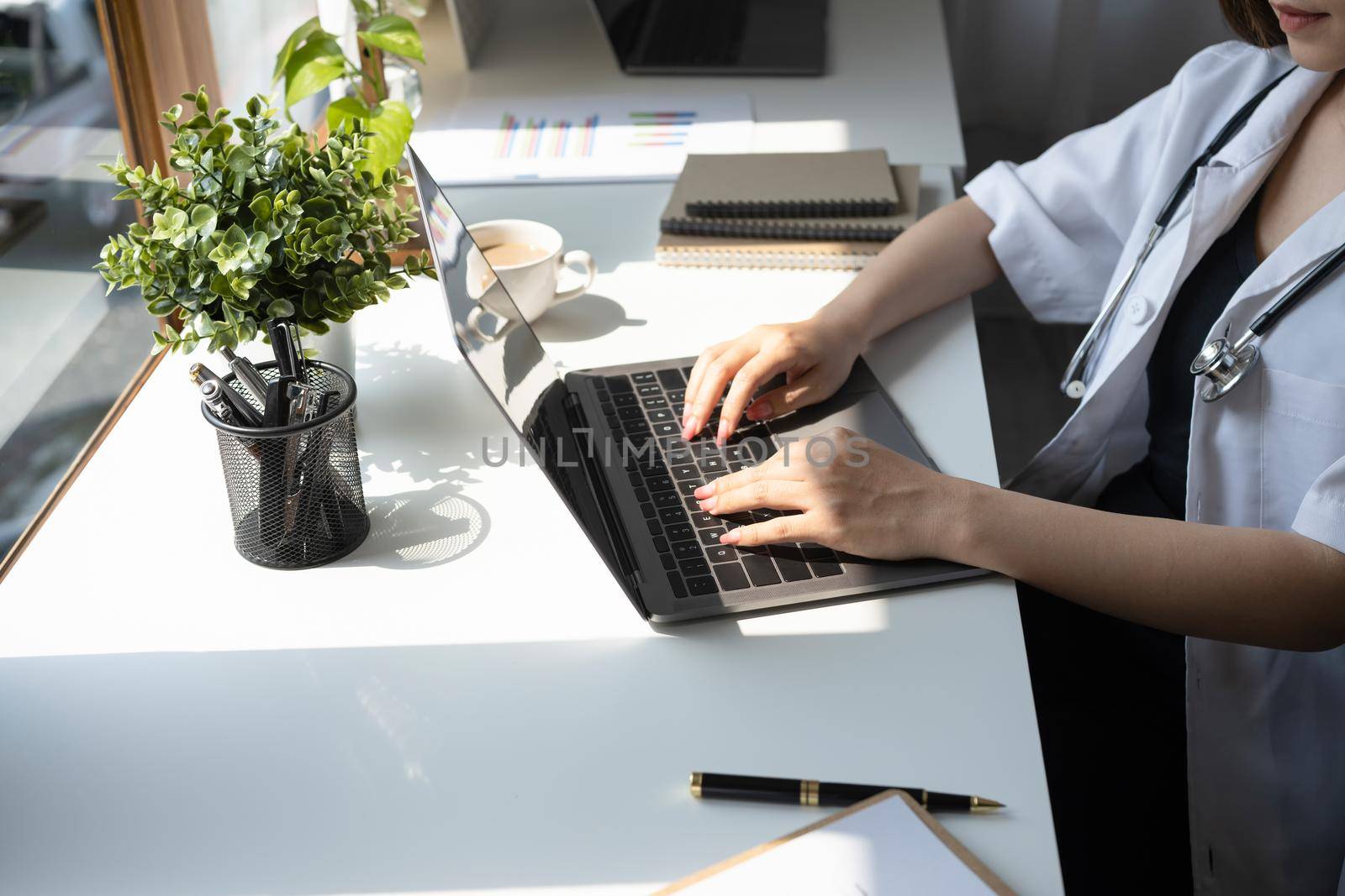 Female doctor in white uniform is working with laptop and documents in medical office.