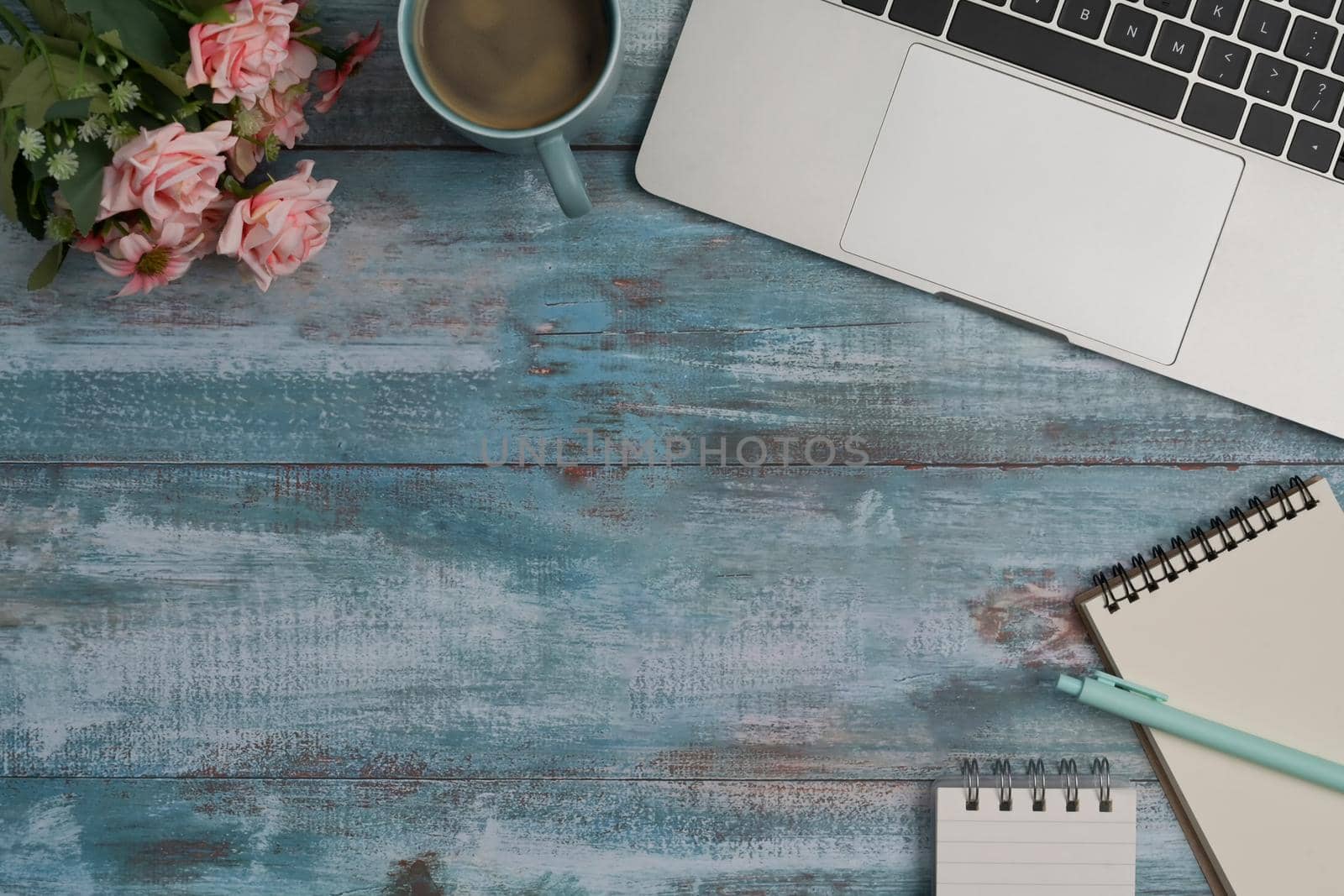 Stylish workplace with laptop computer, notepad, coffee cup and rose flowers on blue wooden desk.