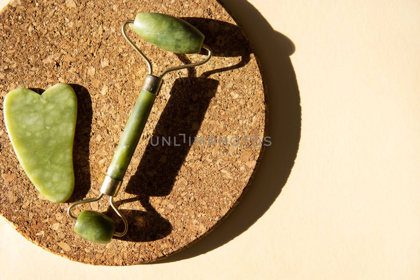 Jade Gua sha scraper and face roller massager on a cork round stand with a monstera leaf. Hard light, shadows, the concept self-care. Facial care. Zero waste. Lifting and toning treatment at home.