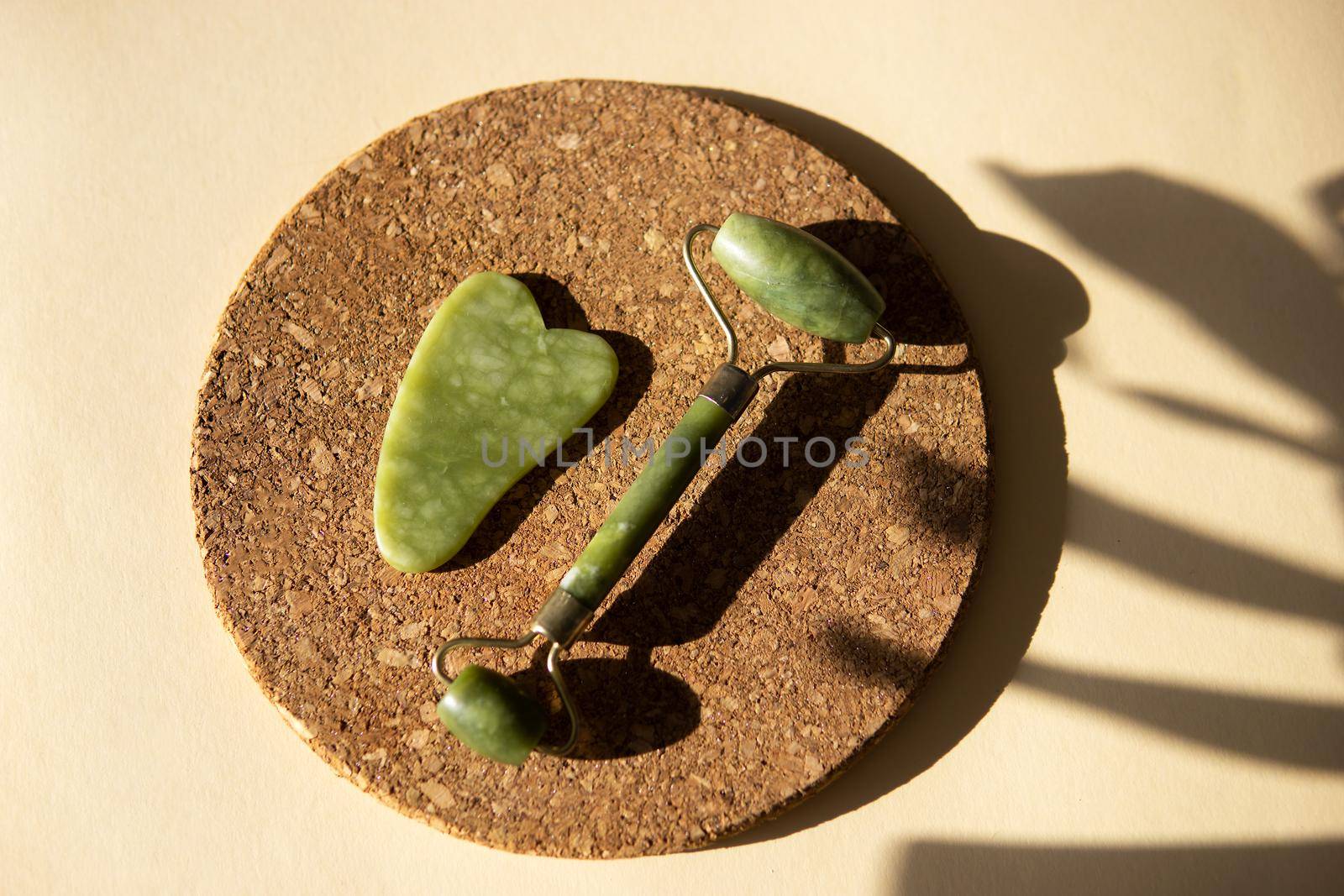 Jade Gua sha scraper and face roller massager on a cork round stand with a monstera leaf. Hard light, shadows, the concept self-care. Facial care. Zero waste. Lifting and toning treatment at home.