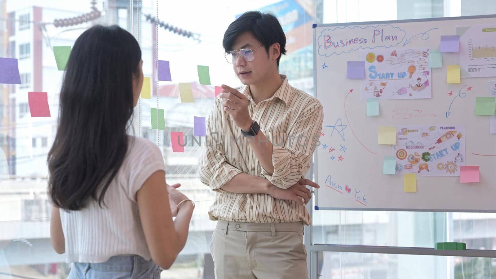 Young creative woman standing near flip chart with her colleague and discussing new marketing strategy together. by prathanchorruangsak