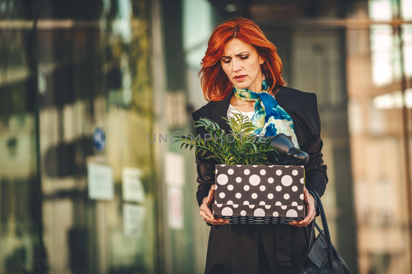 Serious pensive business woman is walking in office district with a box full of her personal belongings from the office just after she got fired. 