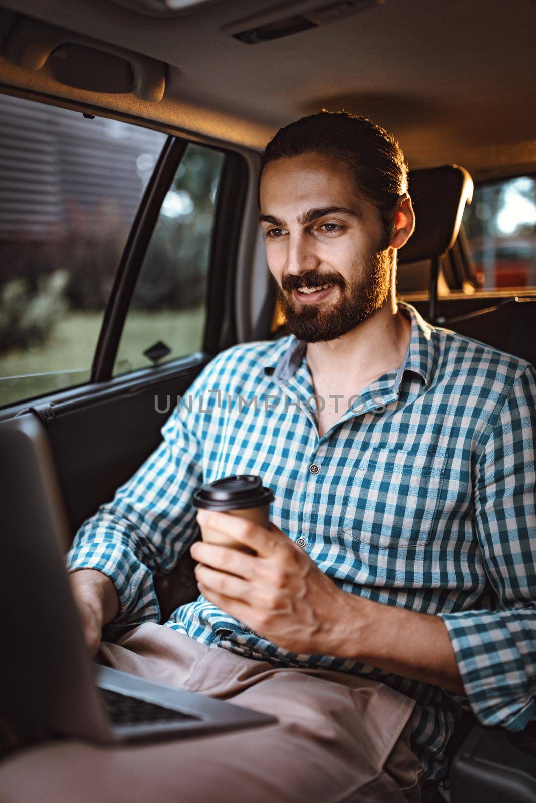 Young confident businessman is holding a cup of coffee in his hand, drinking and texting on the back seat in car.