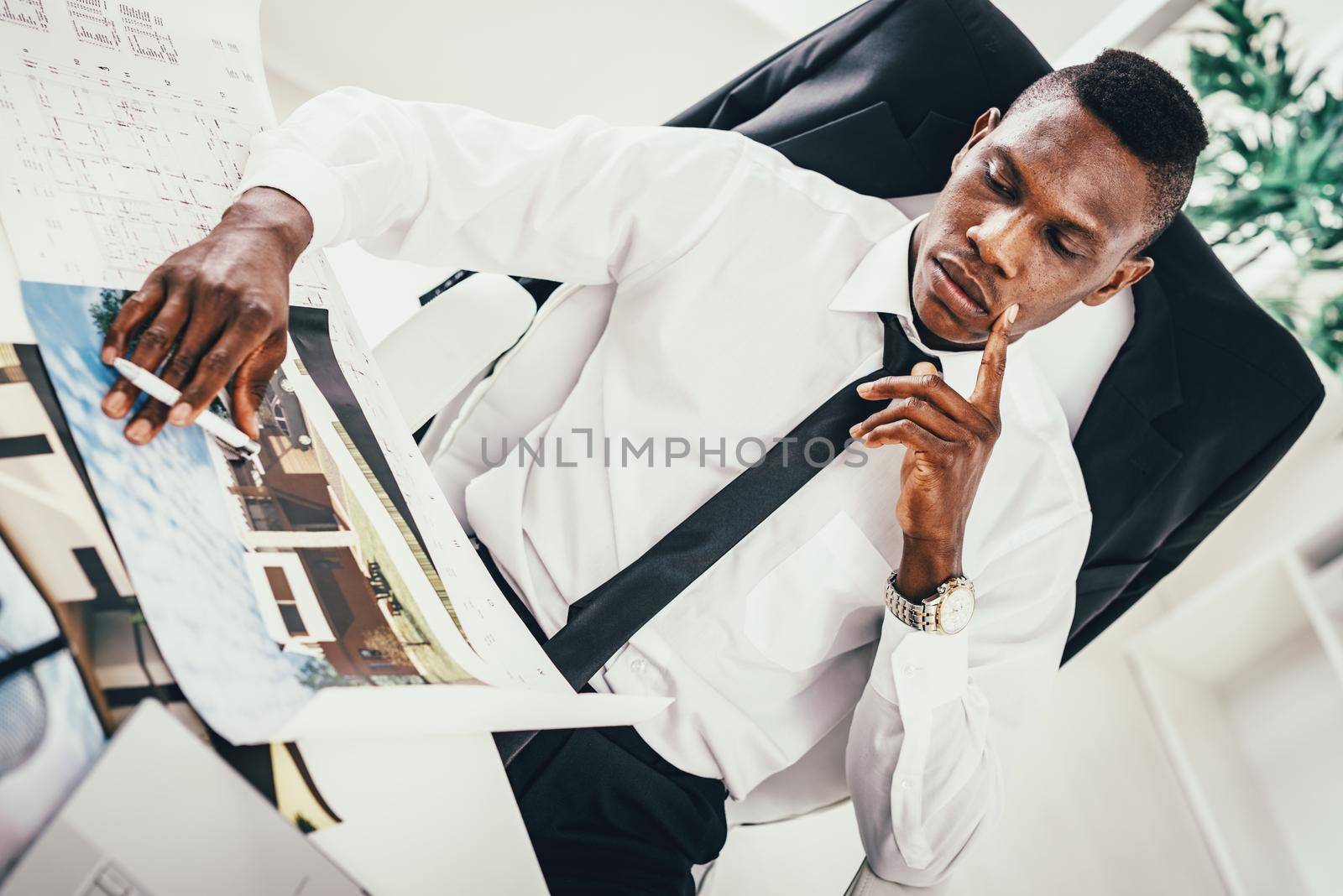 Pensive African architect analyzing blueprint in modern office and planning what to do next.