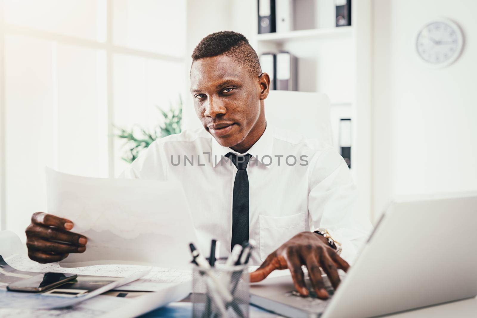 Successful  African businessman looking on paper document and working on laptop in modern office.