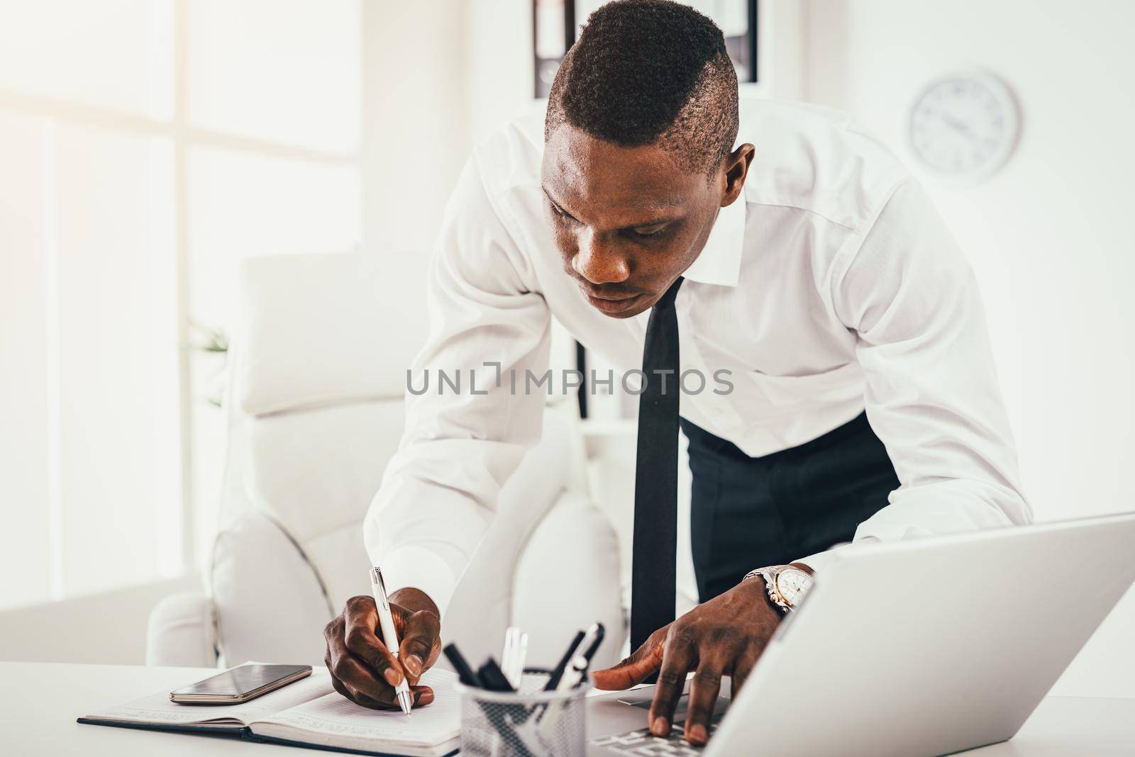 Pensive African businessman is writing notes in notebook and working on laptop in modern office.