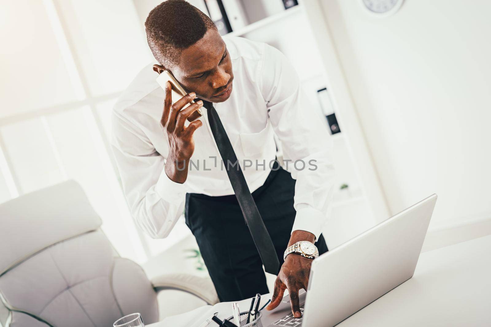 Pensive African businessman talking on smartphone and working at laptop in modern office. 