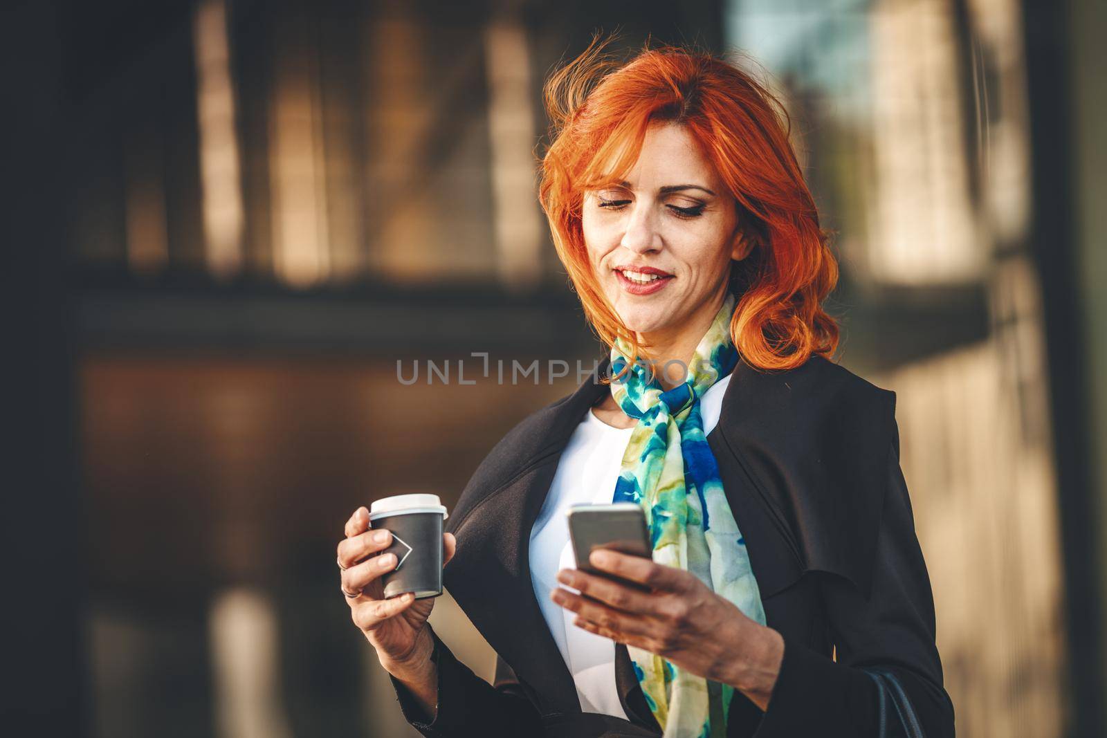 Portrait of a smiling business woman surfing on smartphone on a coffee break in office district at the windy day. She is looking at smartphone with flying hair.