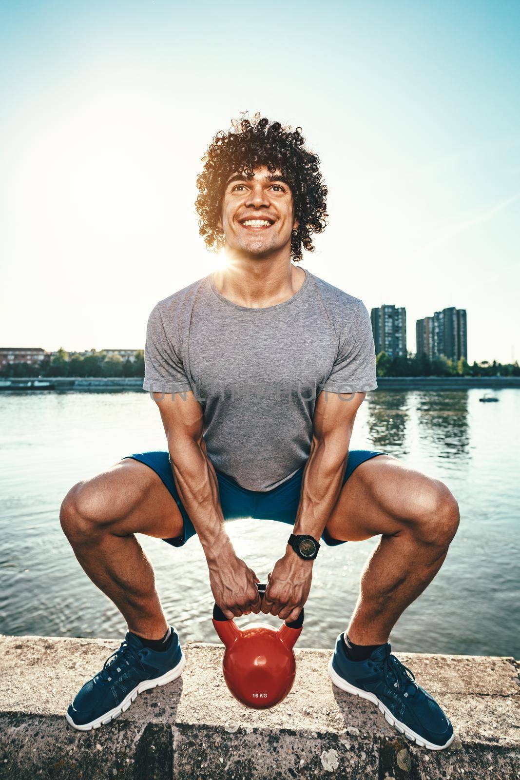 Bodybuilder is crouching and doing strong fit body training with kettlebell on the wall, near the river against blue sky with sunrise light.