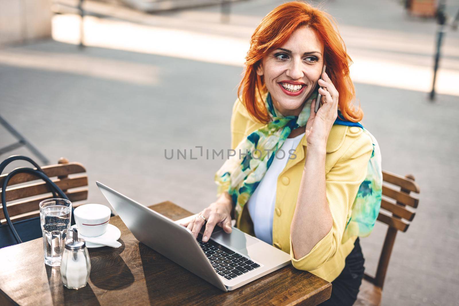 Business woman is working at the laptop and using smartphone on a coffee break in a street cafe.