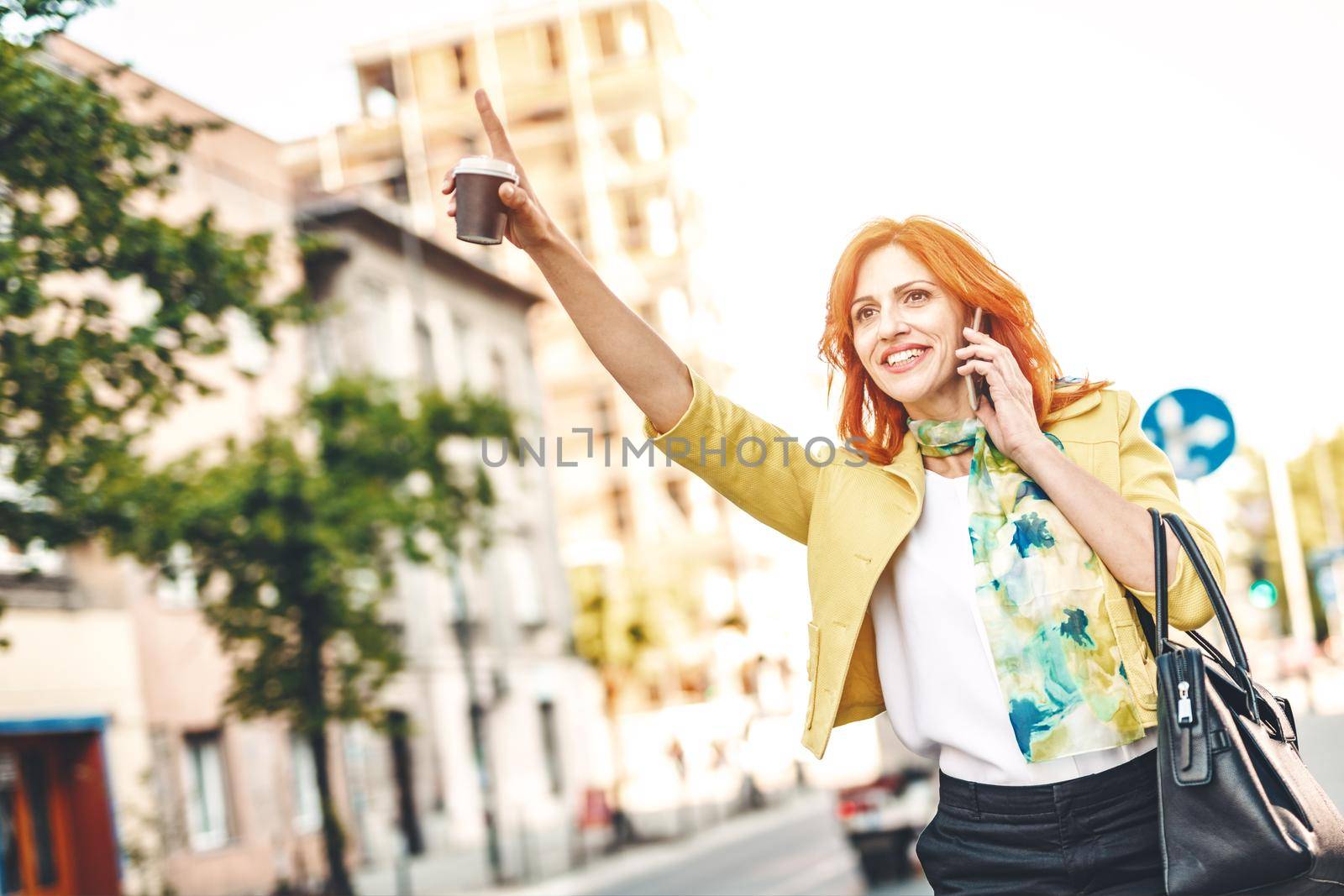 Smiling business woman is standing on the street with smartphone and calling for taxi.