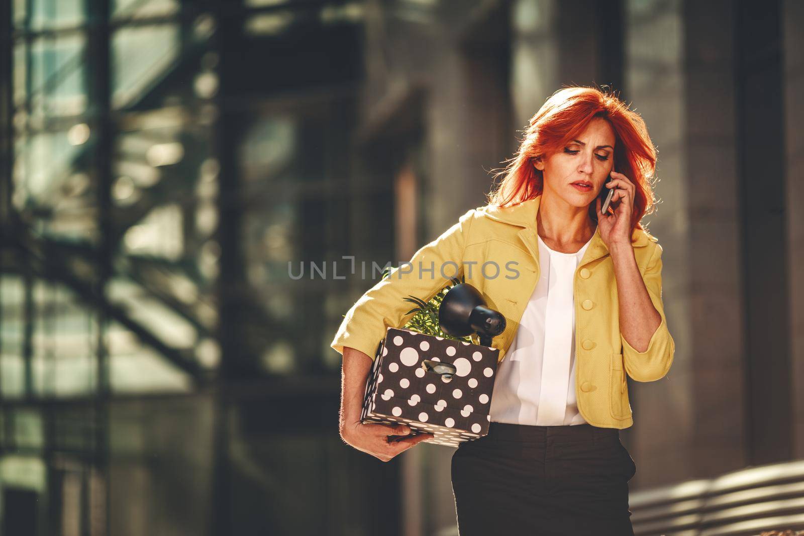 Serious pensive business woman is walking in office district and talking on smartphone with a box full of her personal belongings from the office just after she got fired. 