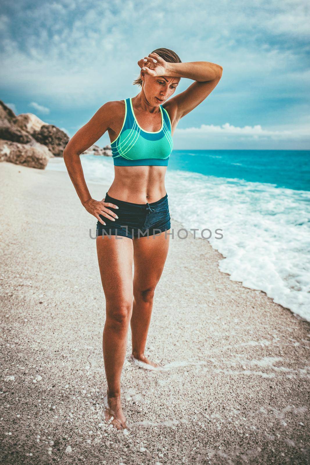 Beautiful young woman relaxing after training on the beach in the end of the day. She is holding her hand on forehead and looking down.