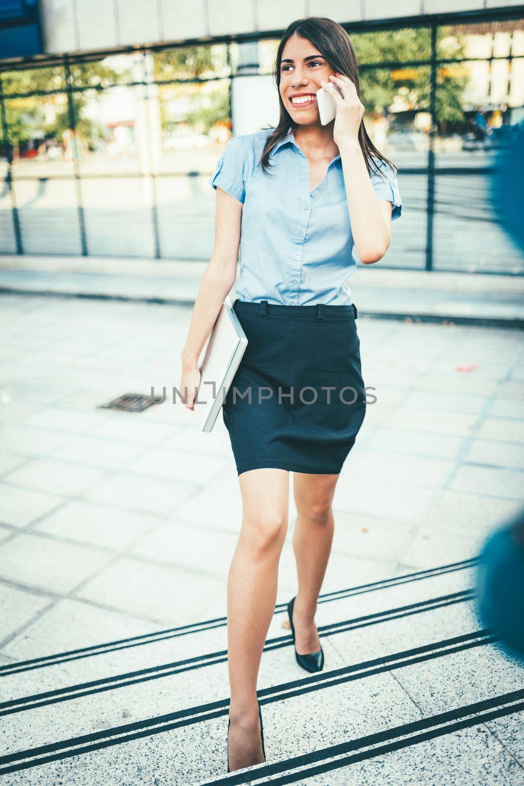 Young beautiful businesswoman walking up the stairs in front of office building, holding a laptop and talking on smartphone.