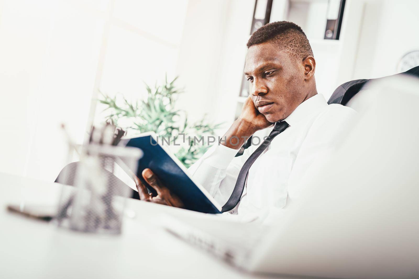 Pensive African businessman working in modern office and reading his notes in notebook and planning what to do next.