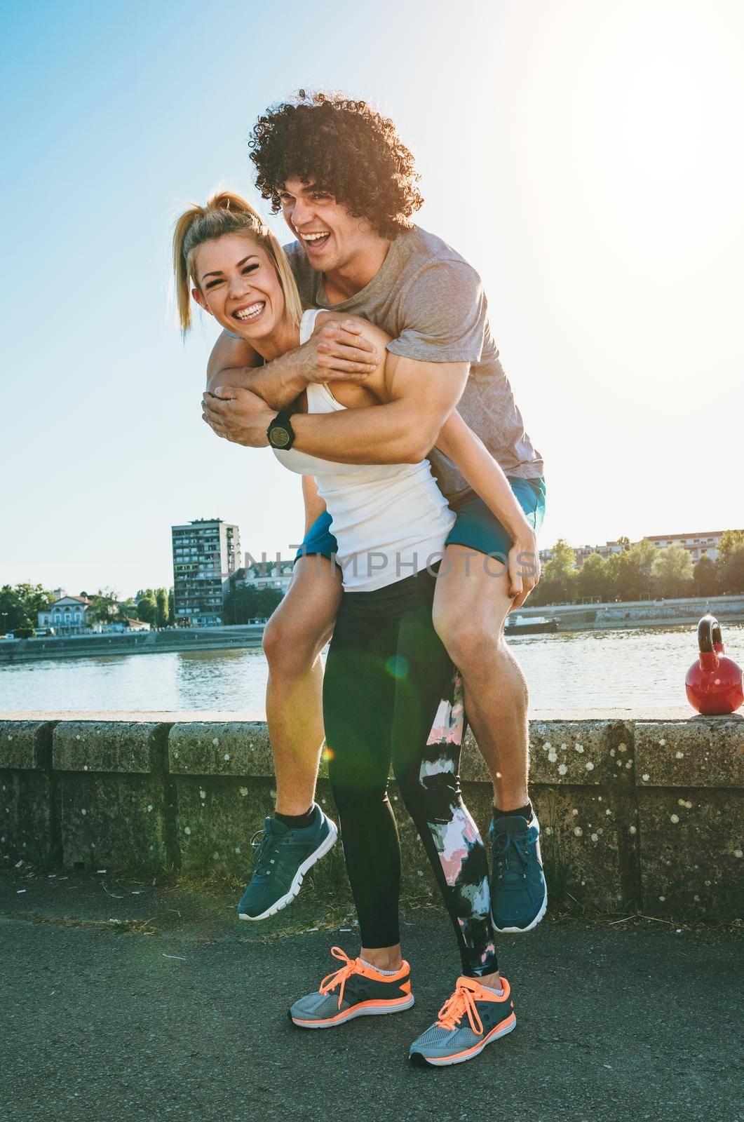Young happy couple runners are having fun after training outdoors by the river, piggybacking in nature against blue sky with sunrise light.