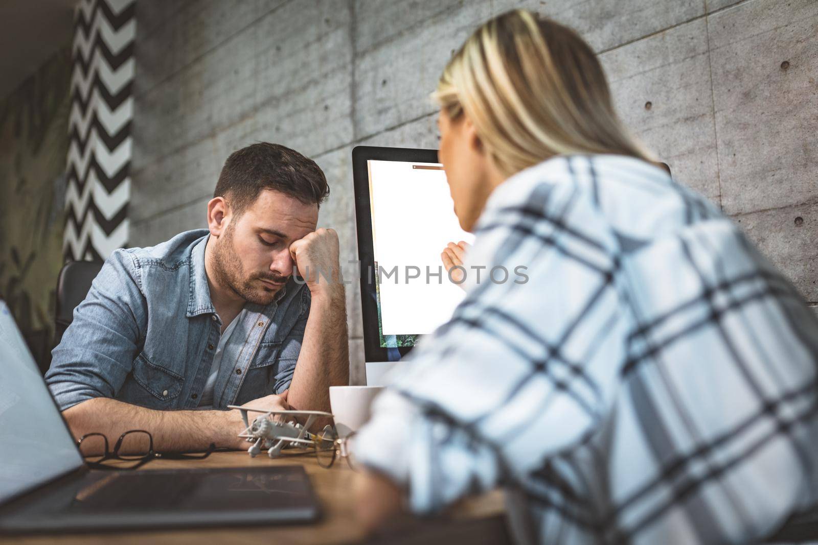 Young handsome frustrated and stressed businessman sitting in the office and complains to his female colleague about the pain in his head.