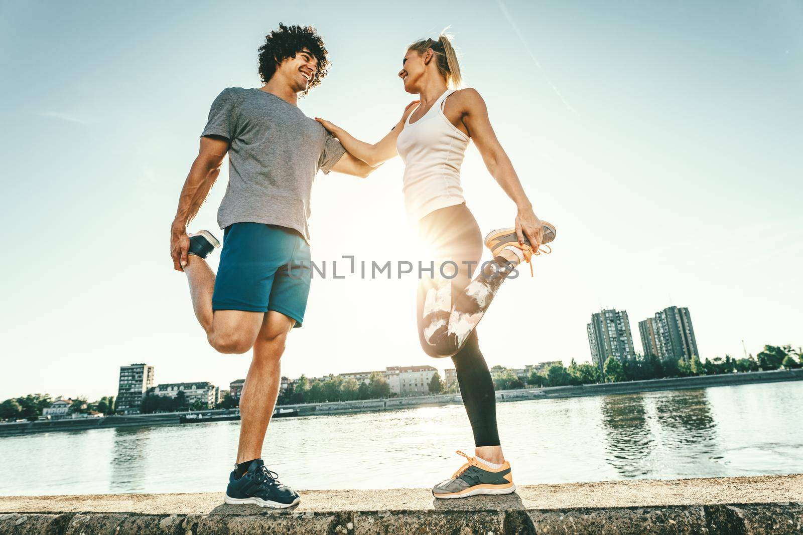 Young happy runners are training outdoors by the river, stretching legs on the wall at sunset.