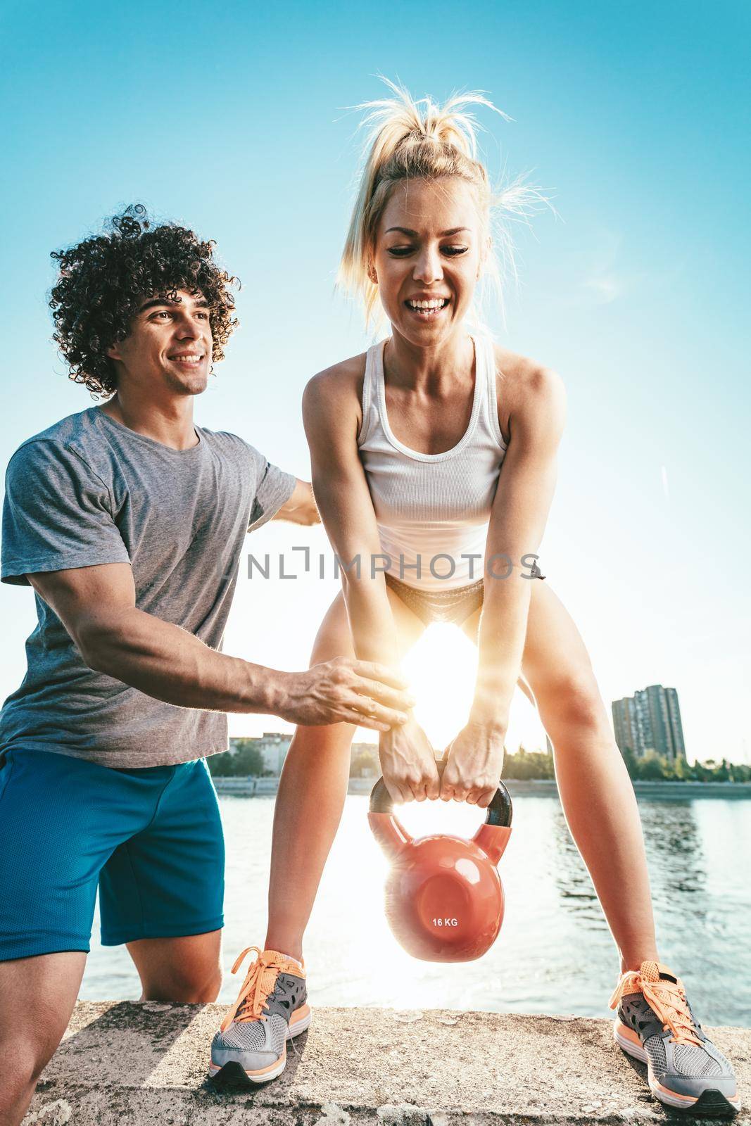 Young fitness couple is doing workout with kettlebell on the wall  by the river in a sunset. The woman is crouching and holding kettlebell, and the man supports her.