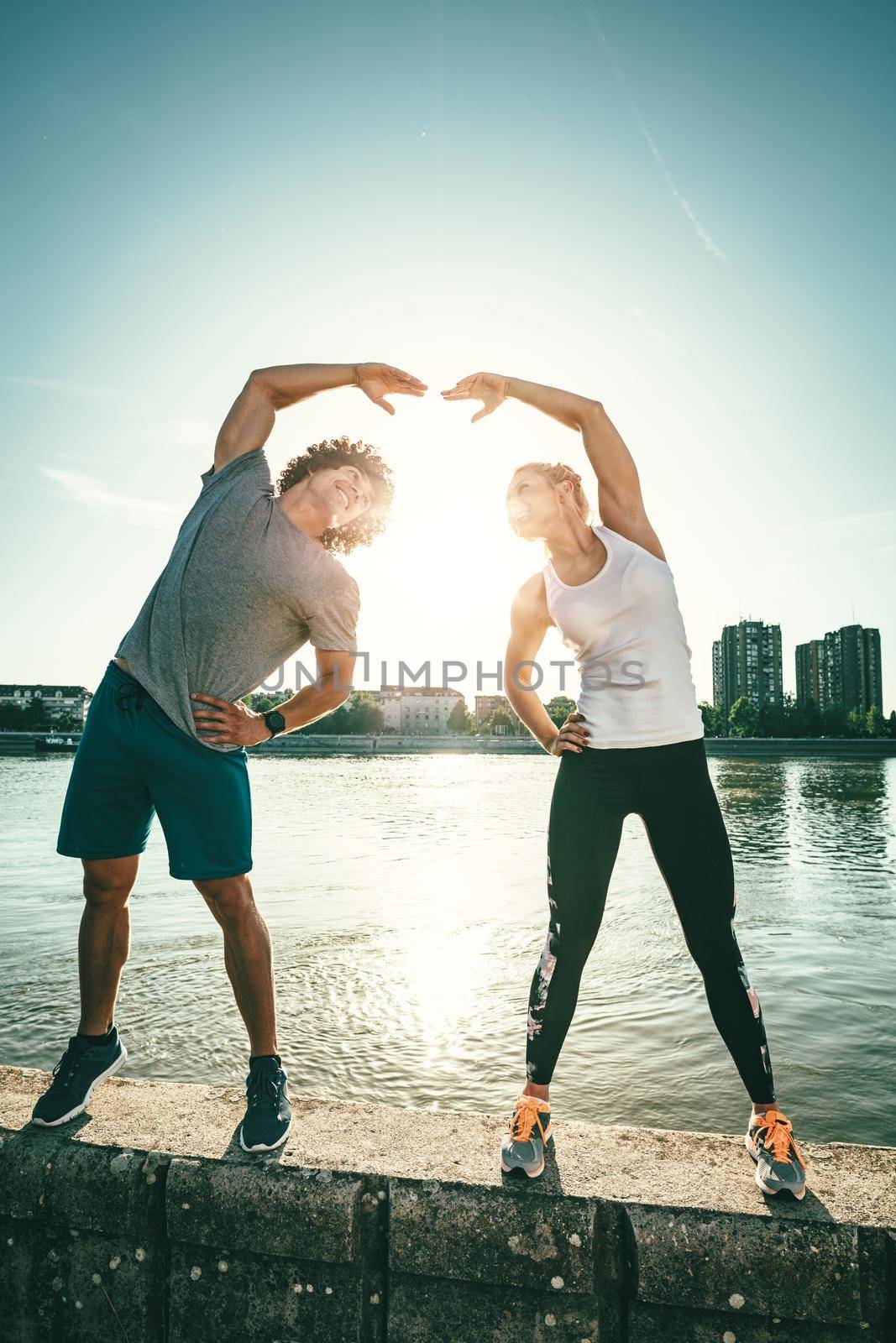 Young happy couple is training outdoors by the river, stretching together on the wall at sunset.