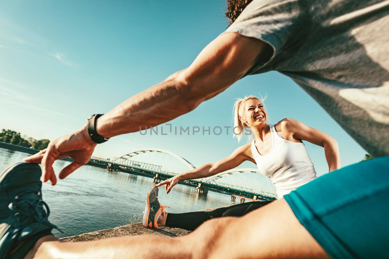 Young happy couple is training outdoors by the river, stretching legs at sunset.