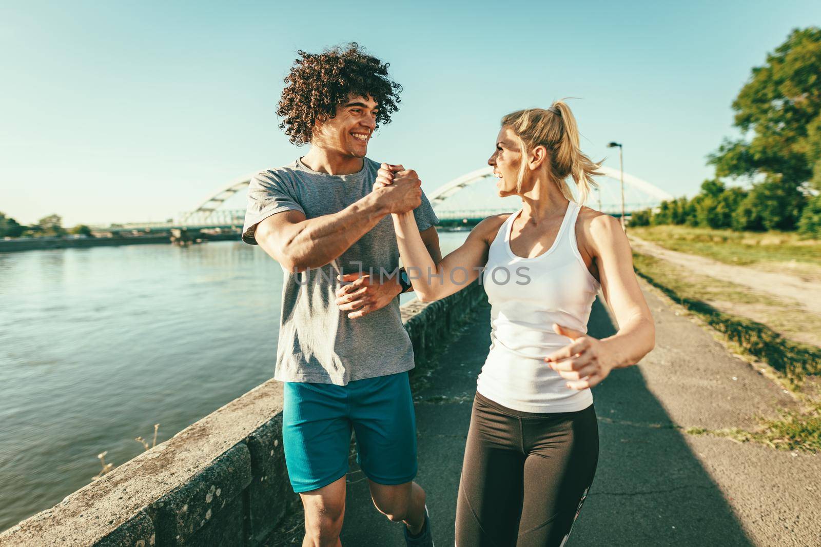 Young happy runners are training outdoors by the river, working out in nature against blue sky with sunrise light.