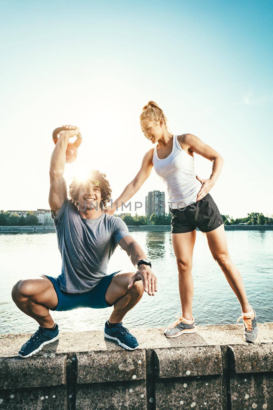 Young fitness couple is doing workout with kettlebell on the wall  by the river in a sunset. The man is crouching and holding kettlebell, and the woman supports him.