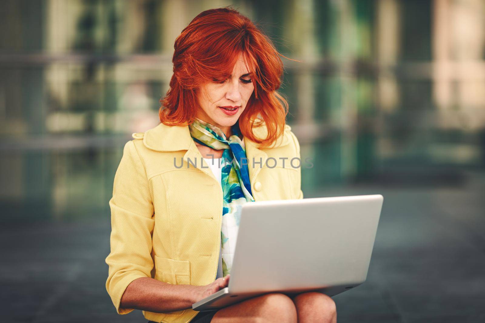 Smiling business woman is sitting on stairs in office district and working at the laptop.