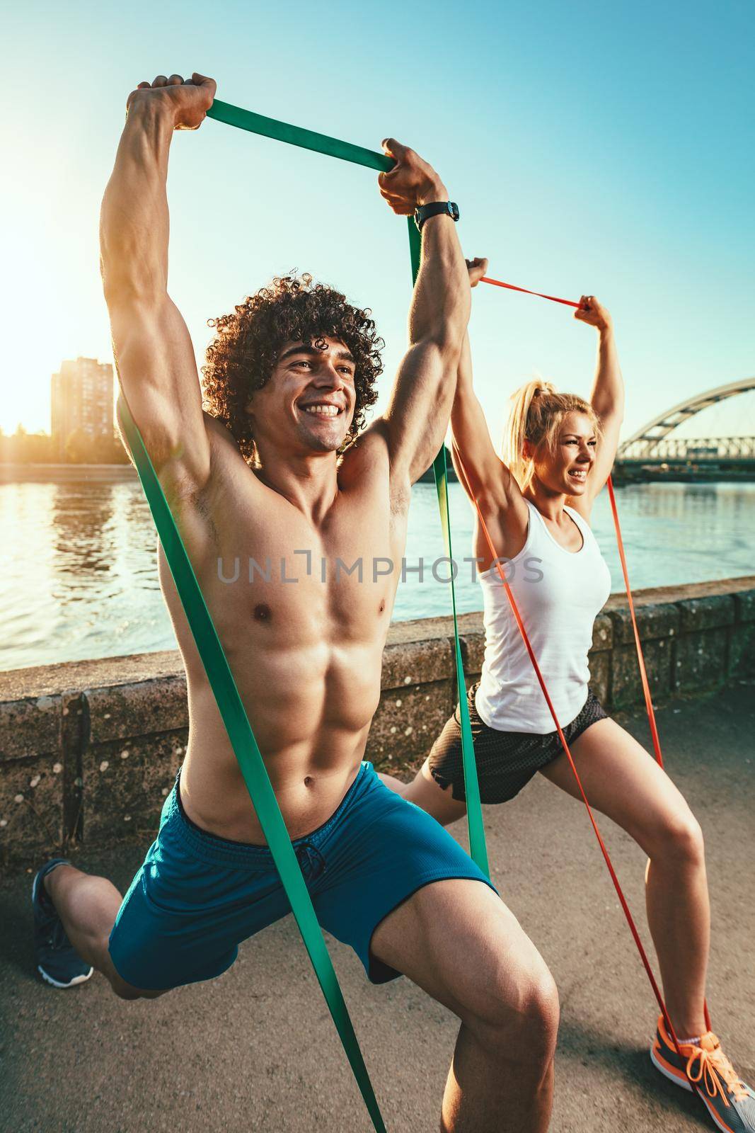 Young fitness couple is doing workout with rubber band by the river in a sunset. 
