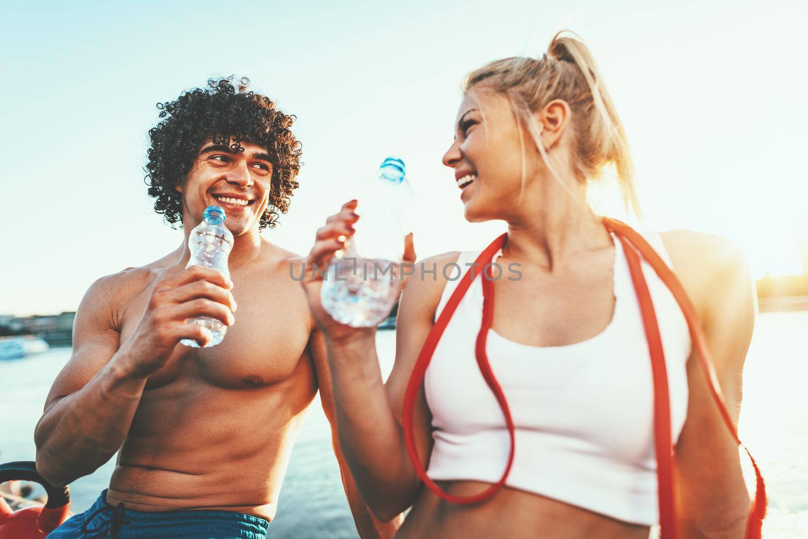 Young fitness couple is resting afther hard training by the river in a sunset drinking water.