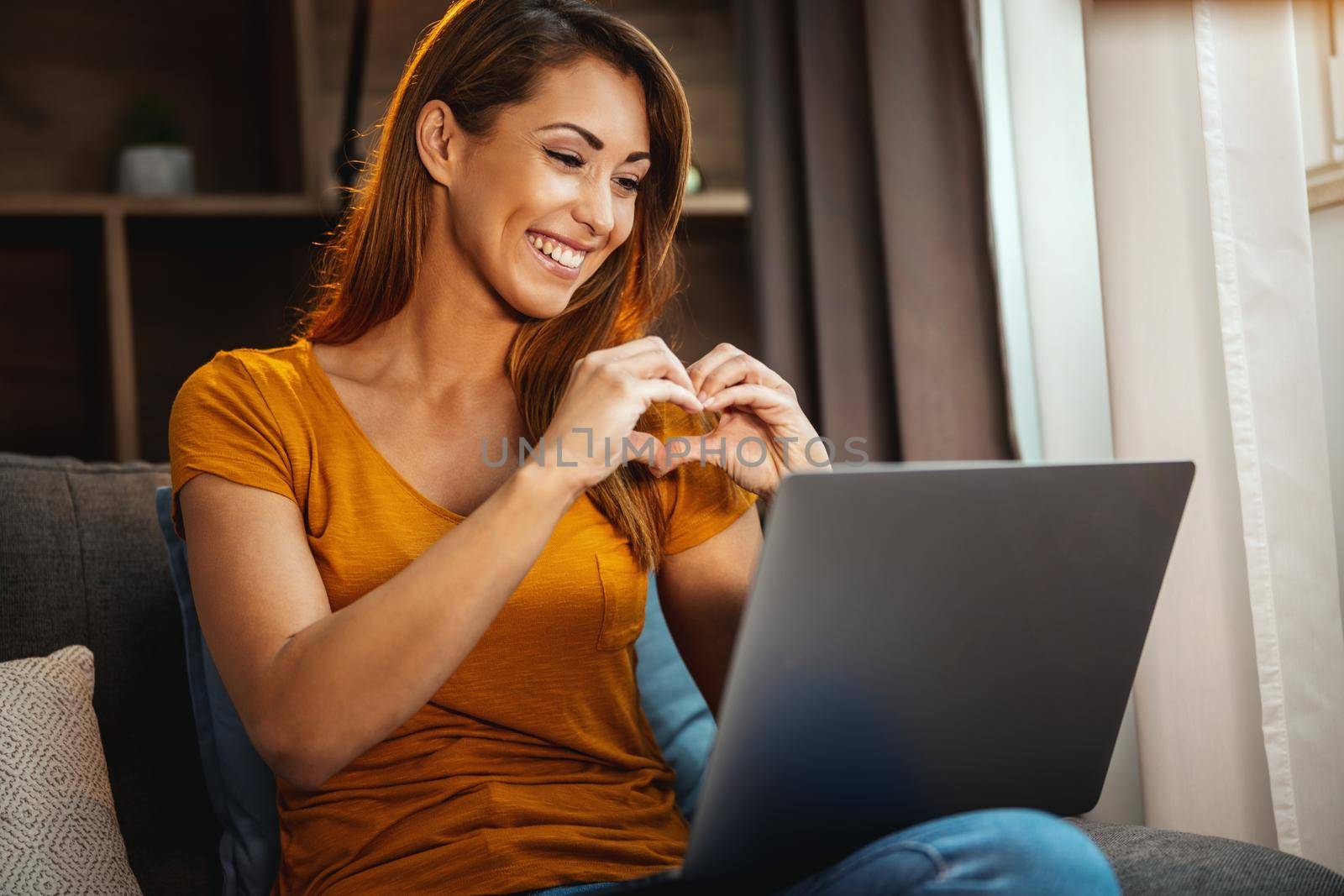 Attractive young woman sitting cross legged on the sofa and using her laptop to make a video chat with someone at home.