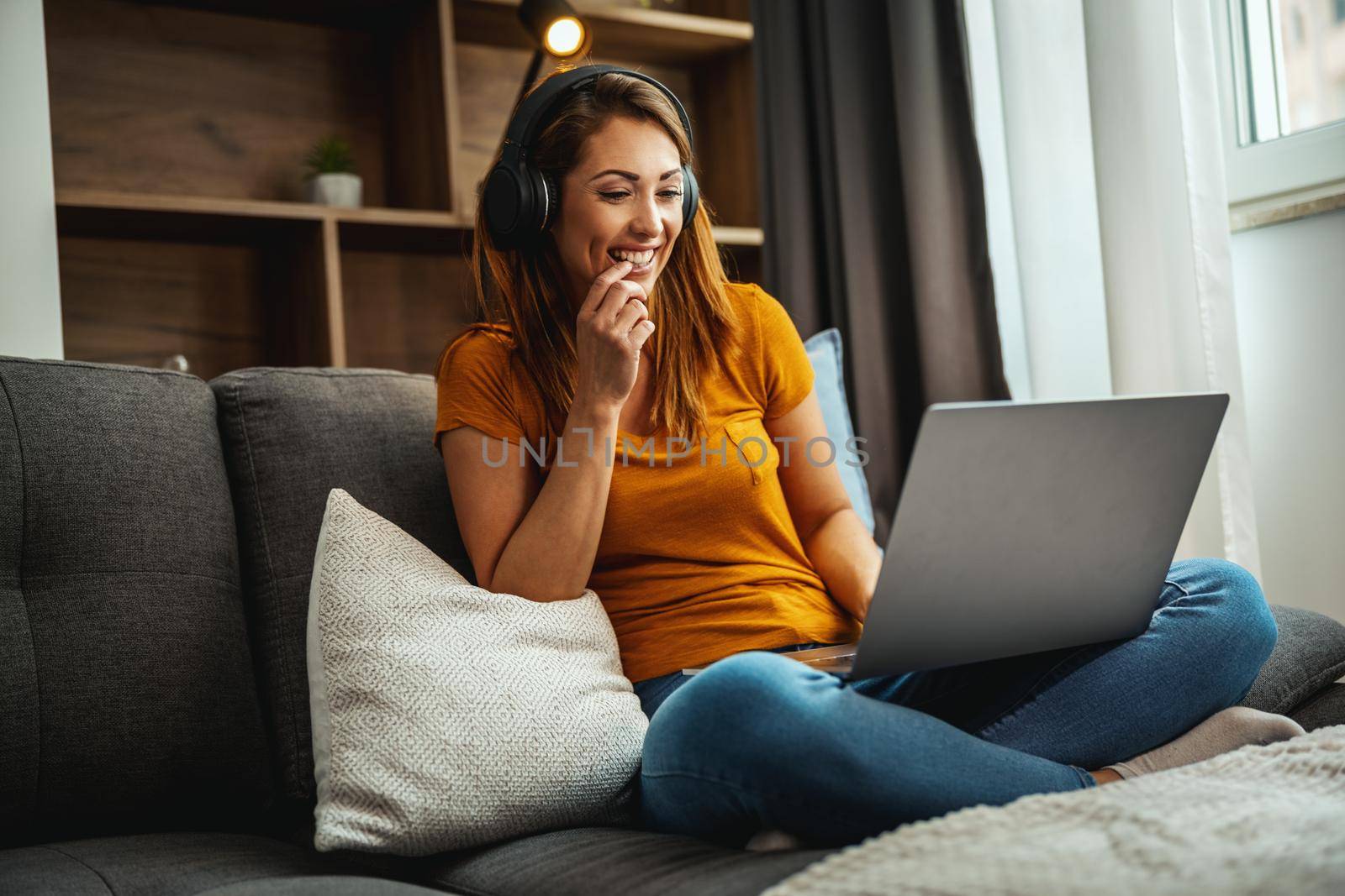 Attractive young woman sitting cross legged on the sofa and using her laptop and headphones to make a video chat with someone at home.