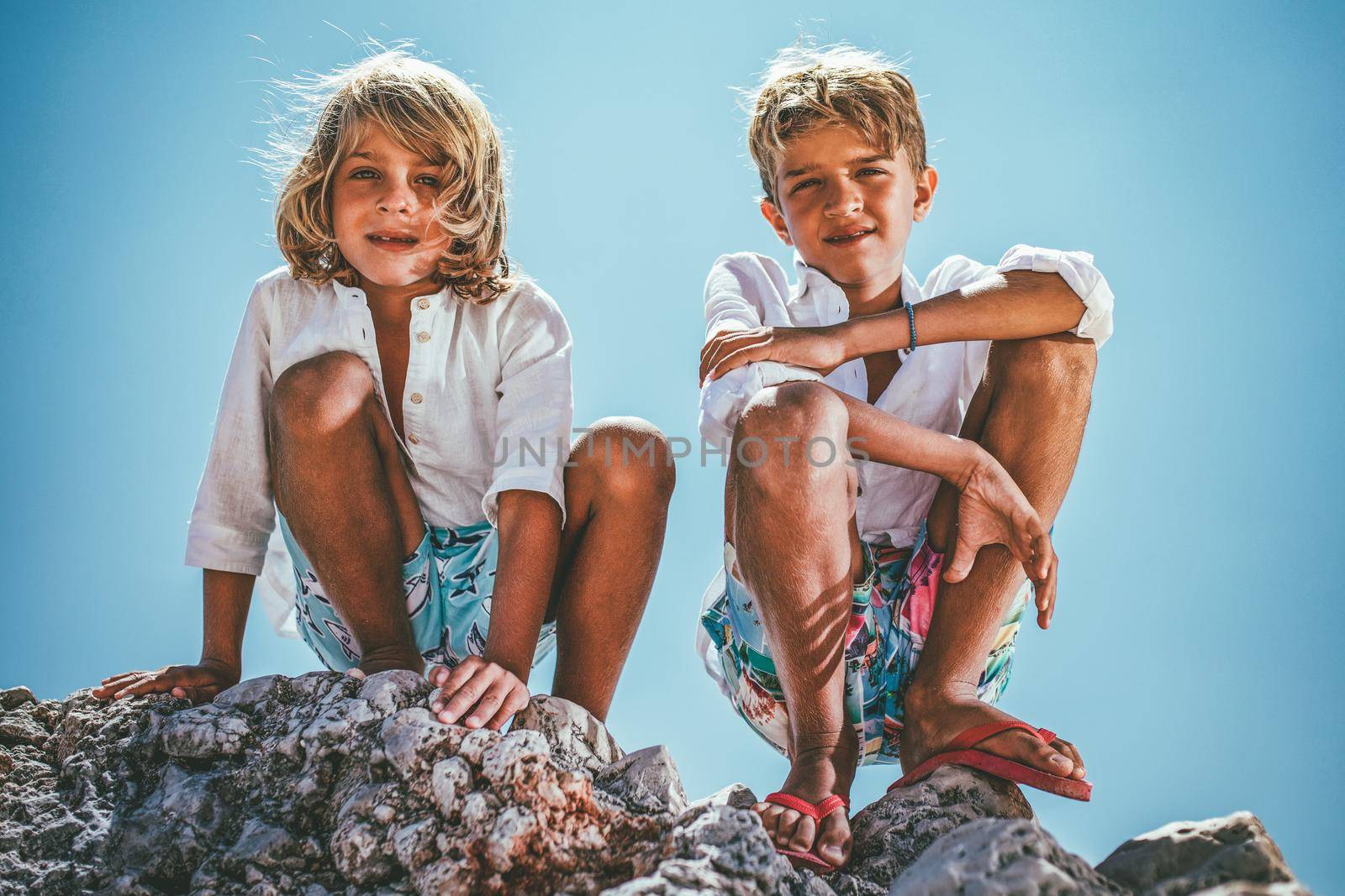 Two cute boys are sitting on the sea rock on the beach and looking down at camera.