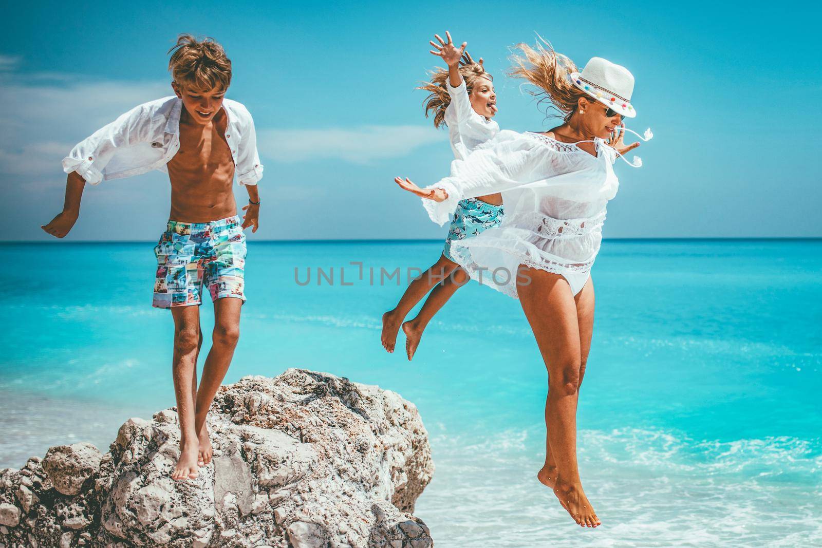 Happy young mother with her little kids having fun at the sunny beach. They are jumping from the rock. 