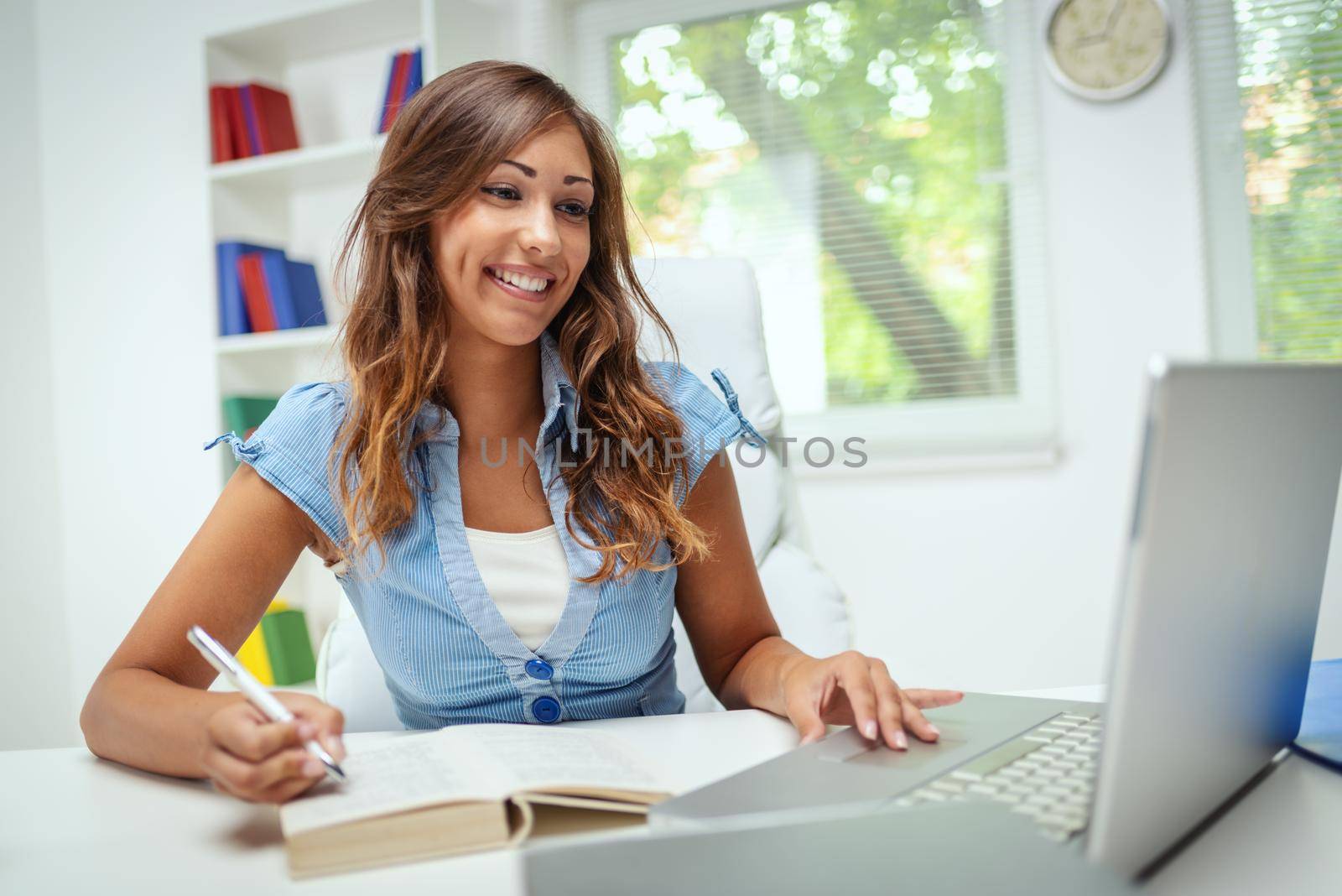 A beautiful smiling young female student is preparing exam and learning lessons in school library, making research on laptop and browse internet.