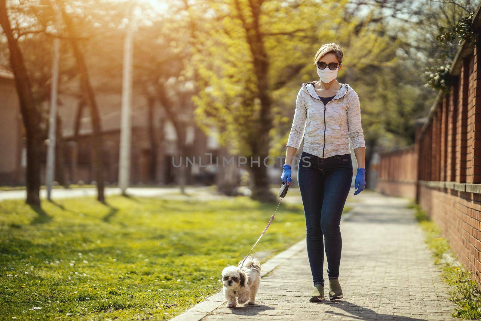 A woman in a medical protective mask is walking along the city street path with her dear cute little Shih Tzu dog during allergy or flu virus outbreak and coronavirus epidemic.