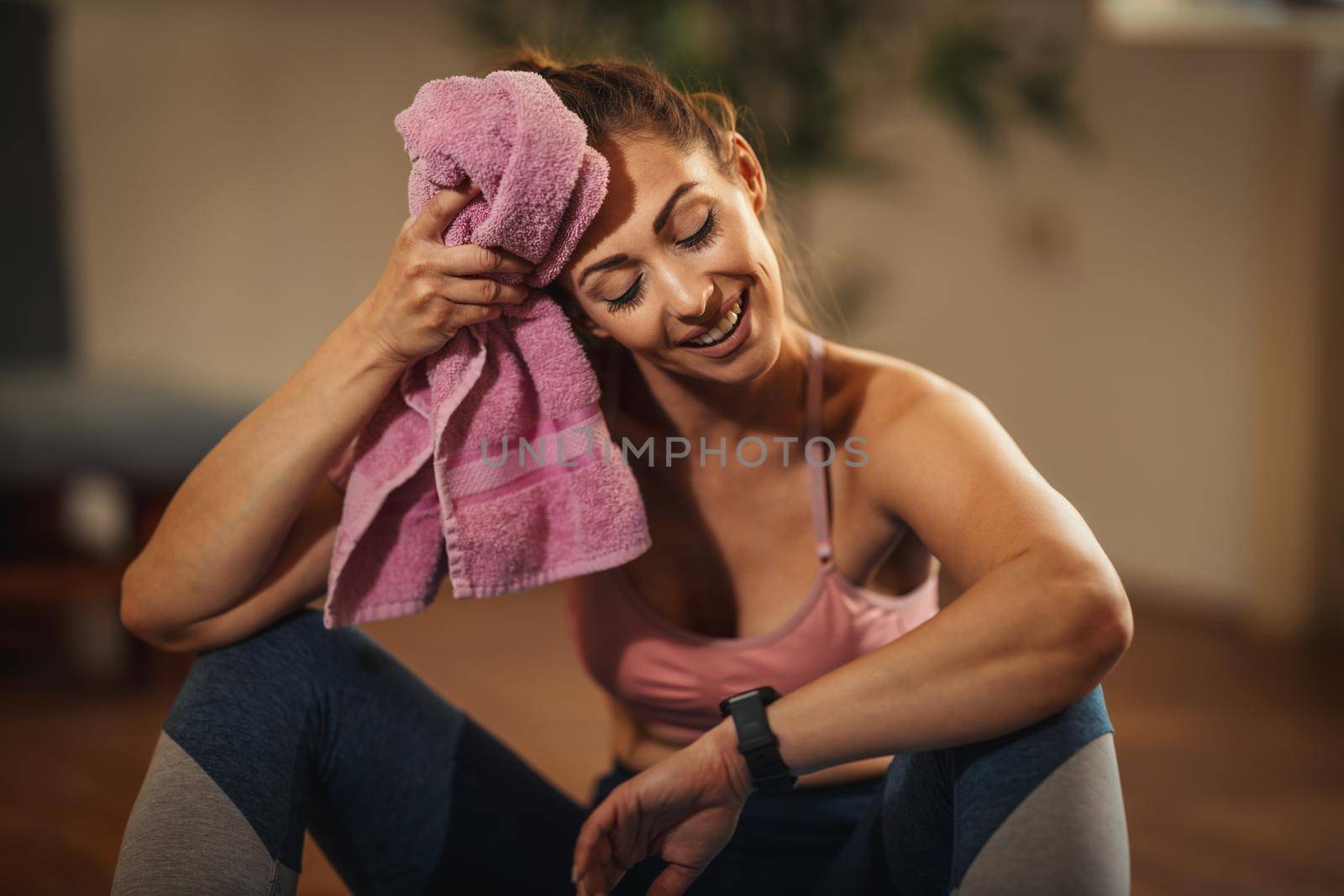 Young woman is resting during sports training in the living room at home. She is enjoying morning sunshine, wiping his forehead with a towel.
