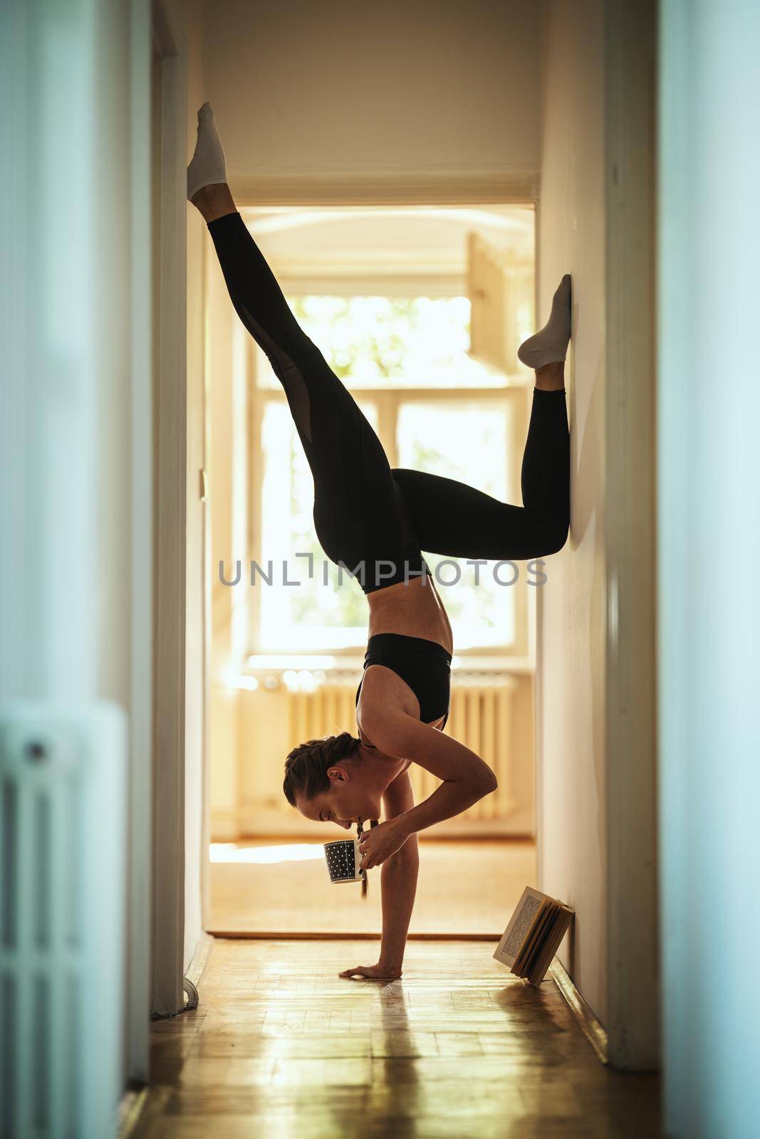 Beautiful fit young woman is drinking coffee and reading book during standing on one hands doing yoga pose.