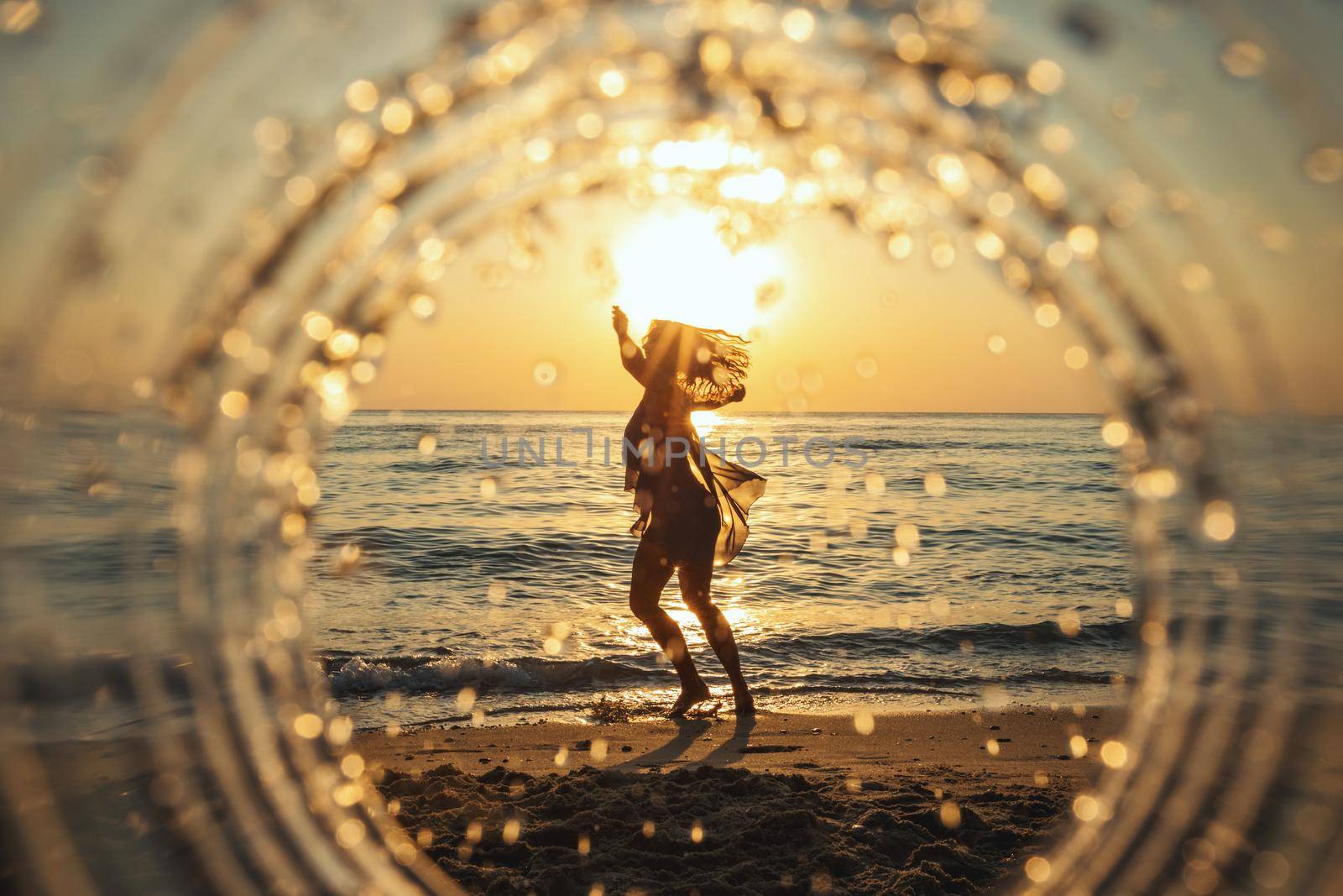 A beautiful creative composition of a sea landscape shot through a circle focus showing a young woman who is dancing on the beach in sunset.